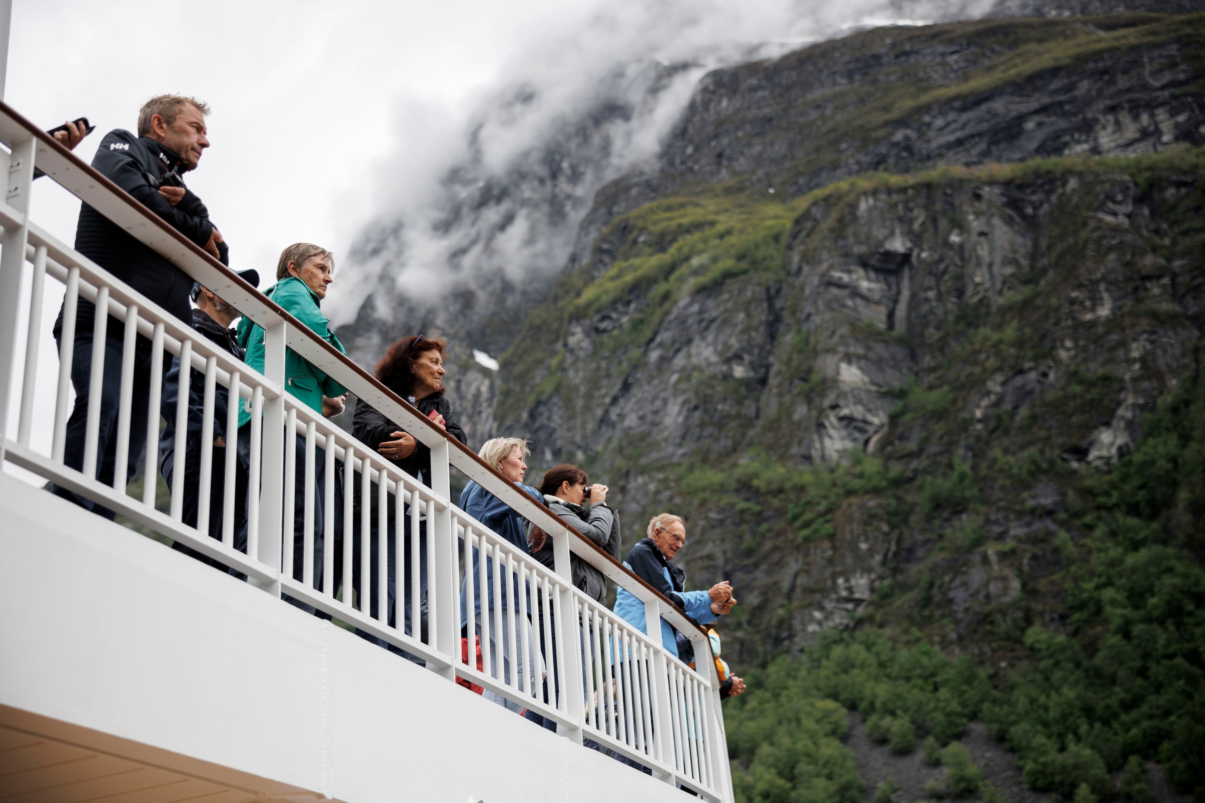 Cruise passengers enjoying panoramic views of Geirangerfjord's towering cliffs and mist-covered mountains from an outdoor deck in Norway