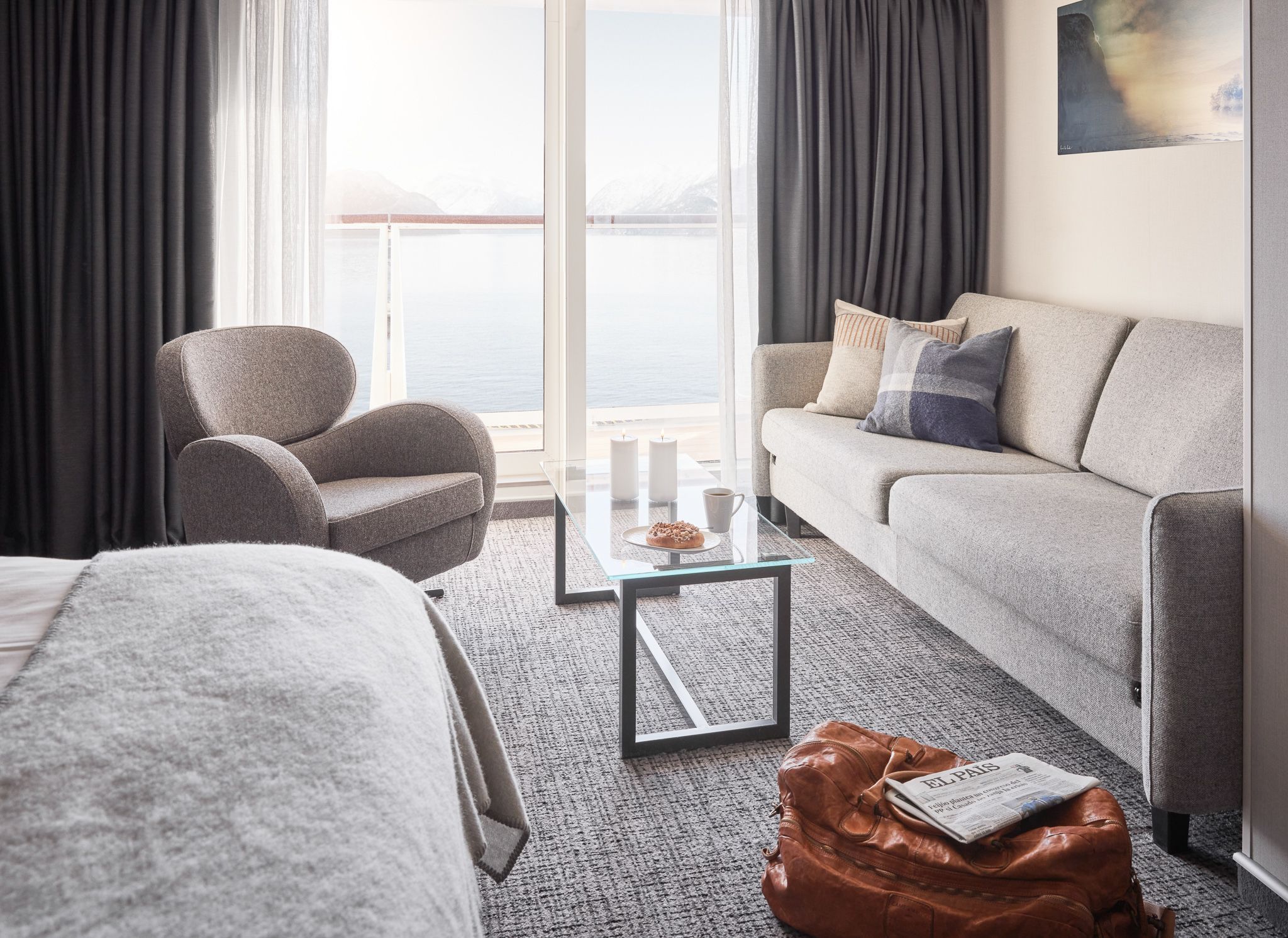 Balcony Junior Suite lounge with a sofa bed, armchair, coffee table, floor-to-ceiling windows, and a private balcony overlooking the sea.