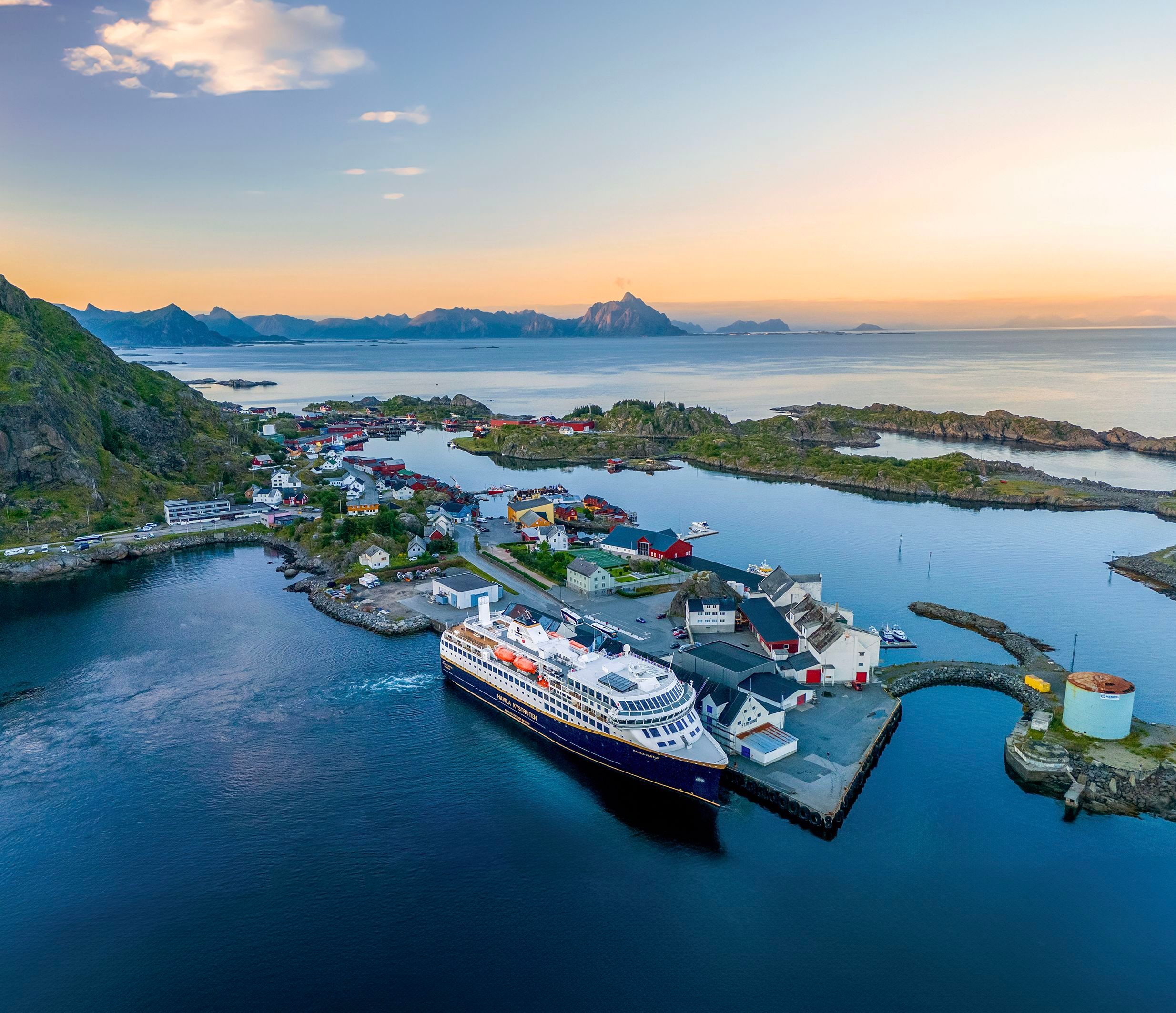 Flyfoto av et Havila Voyages-skip som ligger til kai i Svolvær i Lofoten, omgitt av fargerike sjøhus og dramatiske kystfjell.