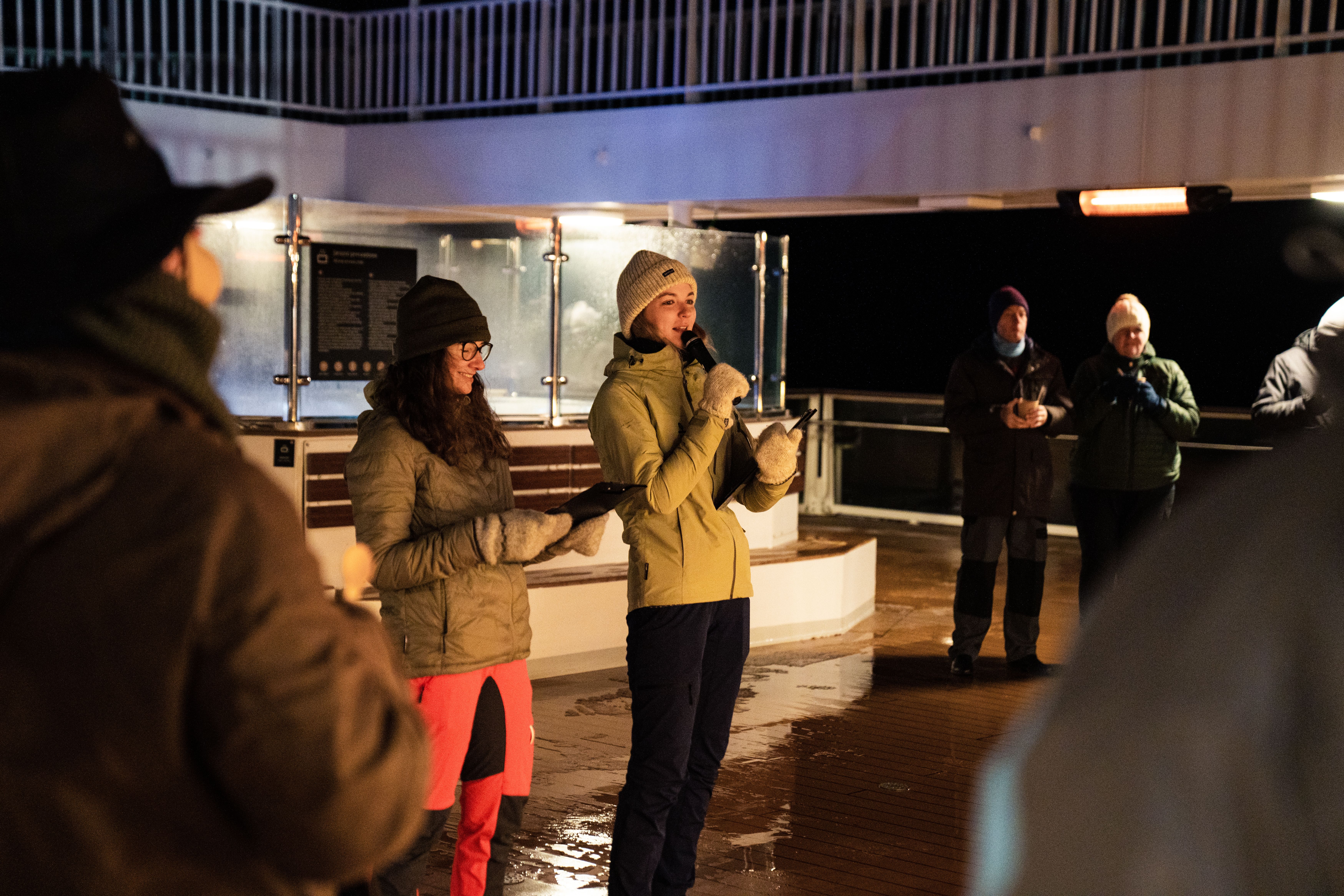 Guests in warm outdoor clothing gathered on deck at night under the lights