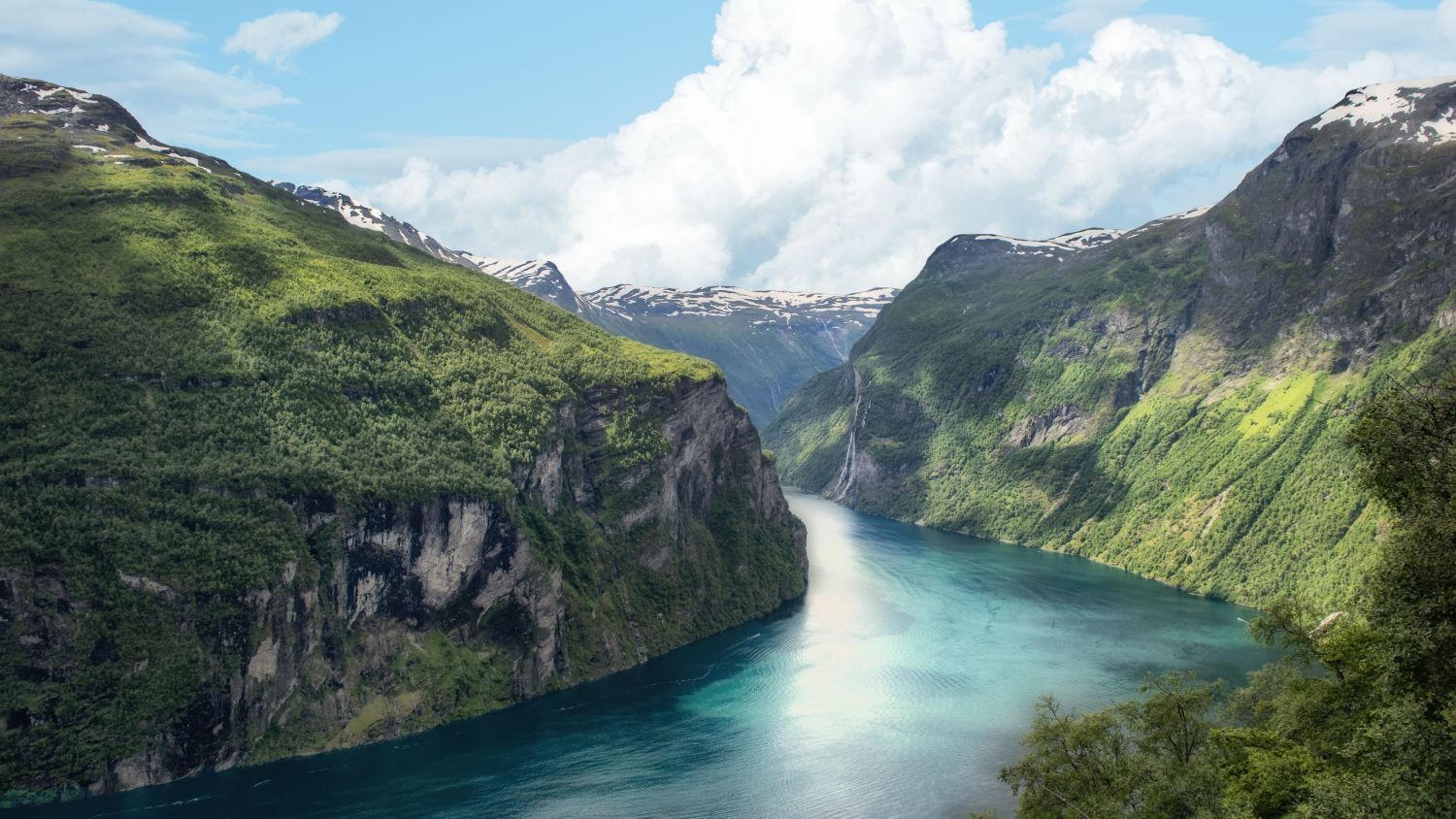 Panoramautsikt over Geirangerfjorden med smaragdgrønt vann, bratte grønne fjell, snødekte topper og fossefall som stuper ned under en klar sommerhimmel