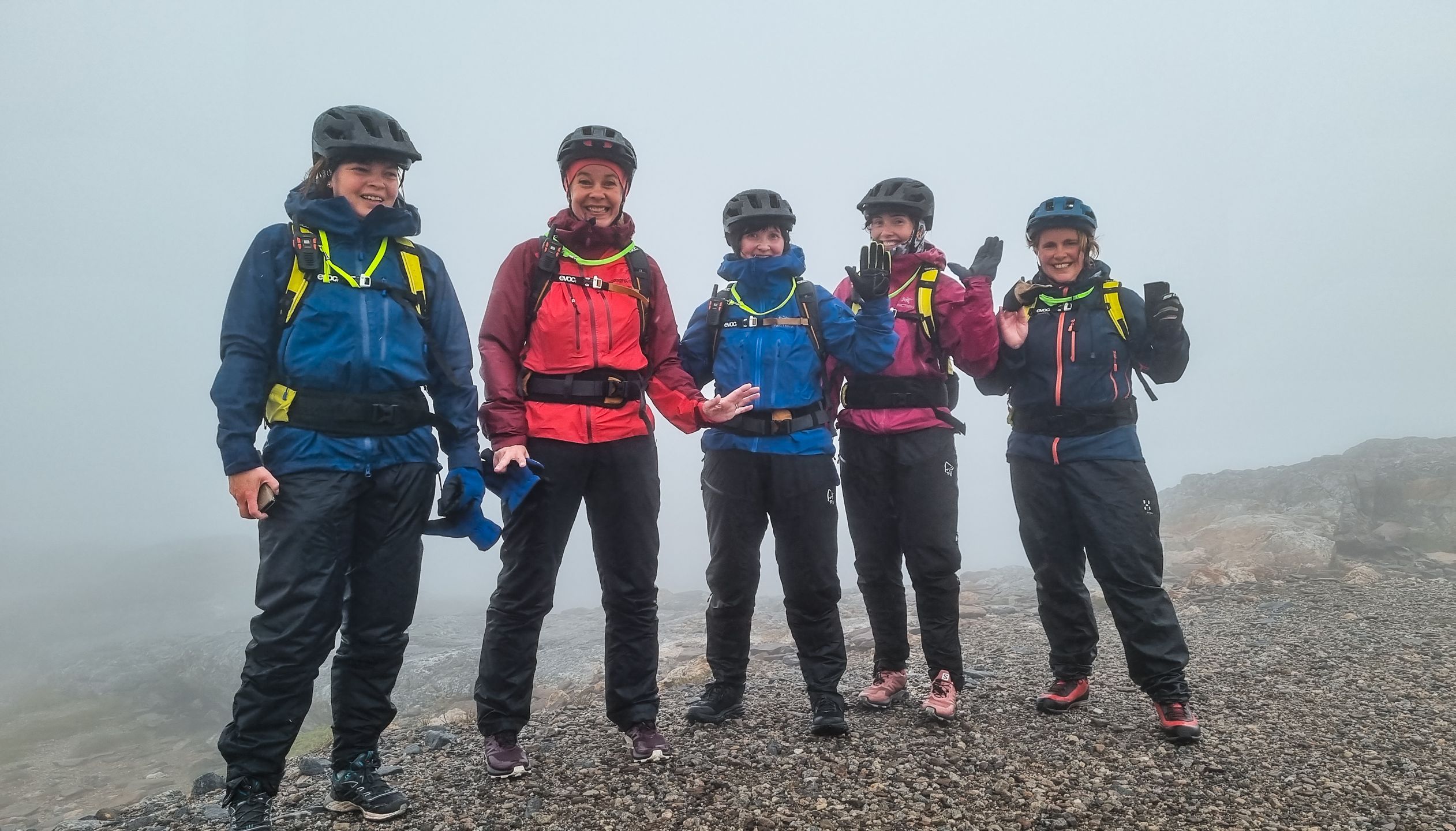 Fem smilende syklister i fargerike regnjakker og hjelmer som vinker på en tåkete fjelltopp under en elsykkeltur i Bodø, Norge