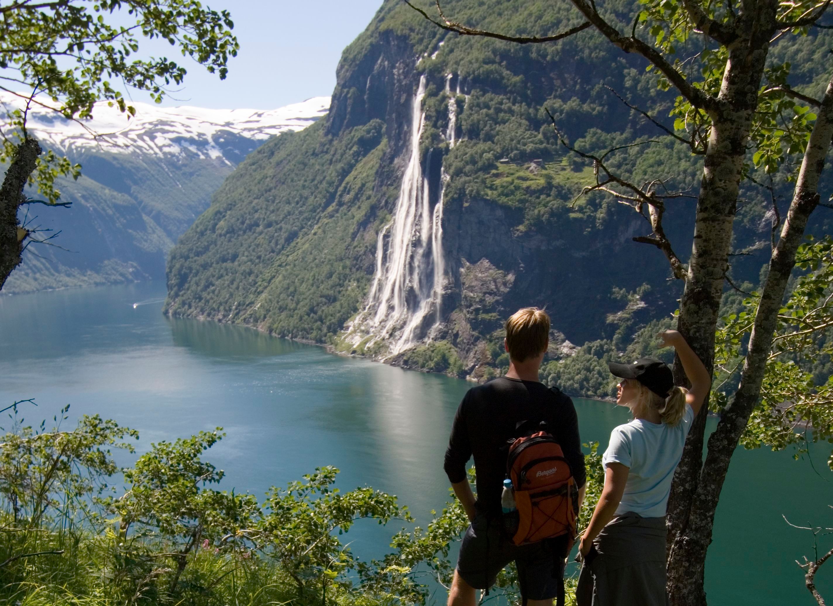 Two hikers admire the Seven Sisters waterfall and the breathtaking landscape of Geirangerfjord, Norway.