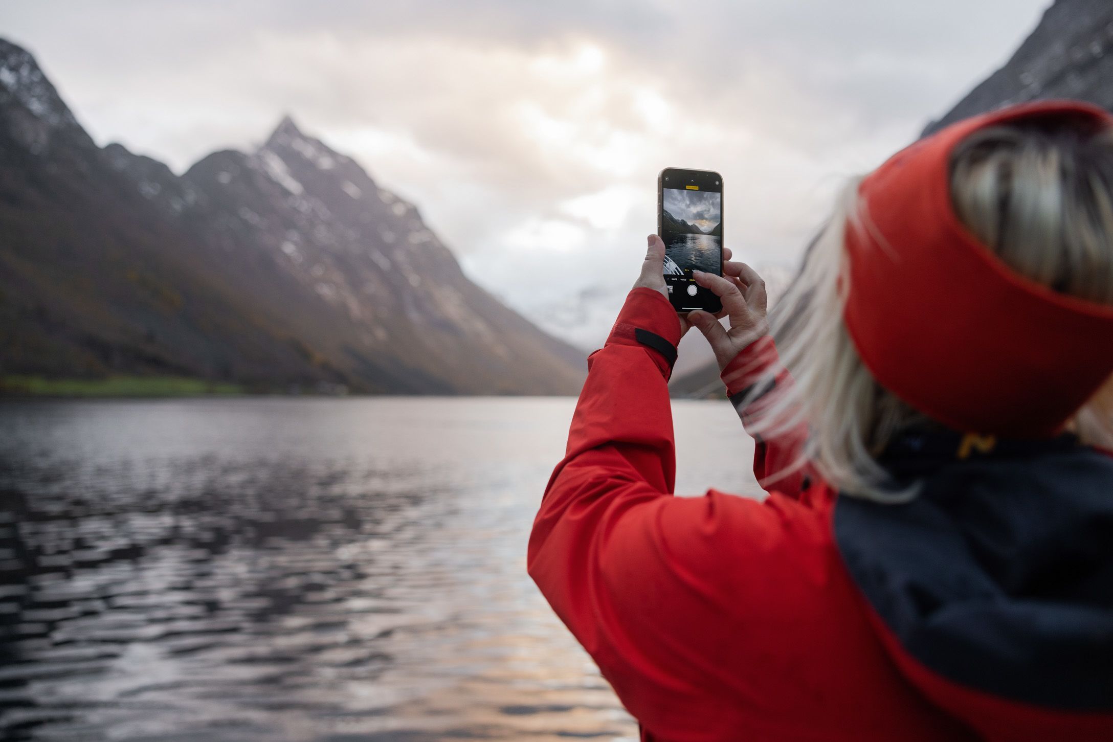 Person in roter Jacke, die mit einem Smartphone einen dramatischen Berggipfel und einen ruhigen Fjord im Hjørundfjord in Norwegen fotografiert