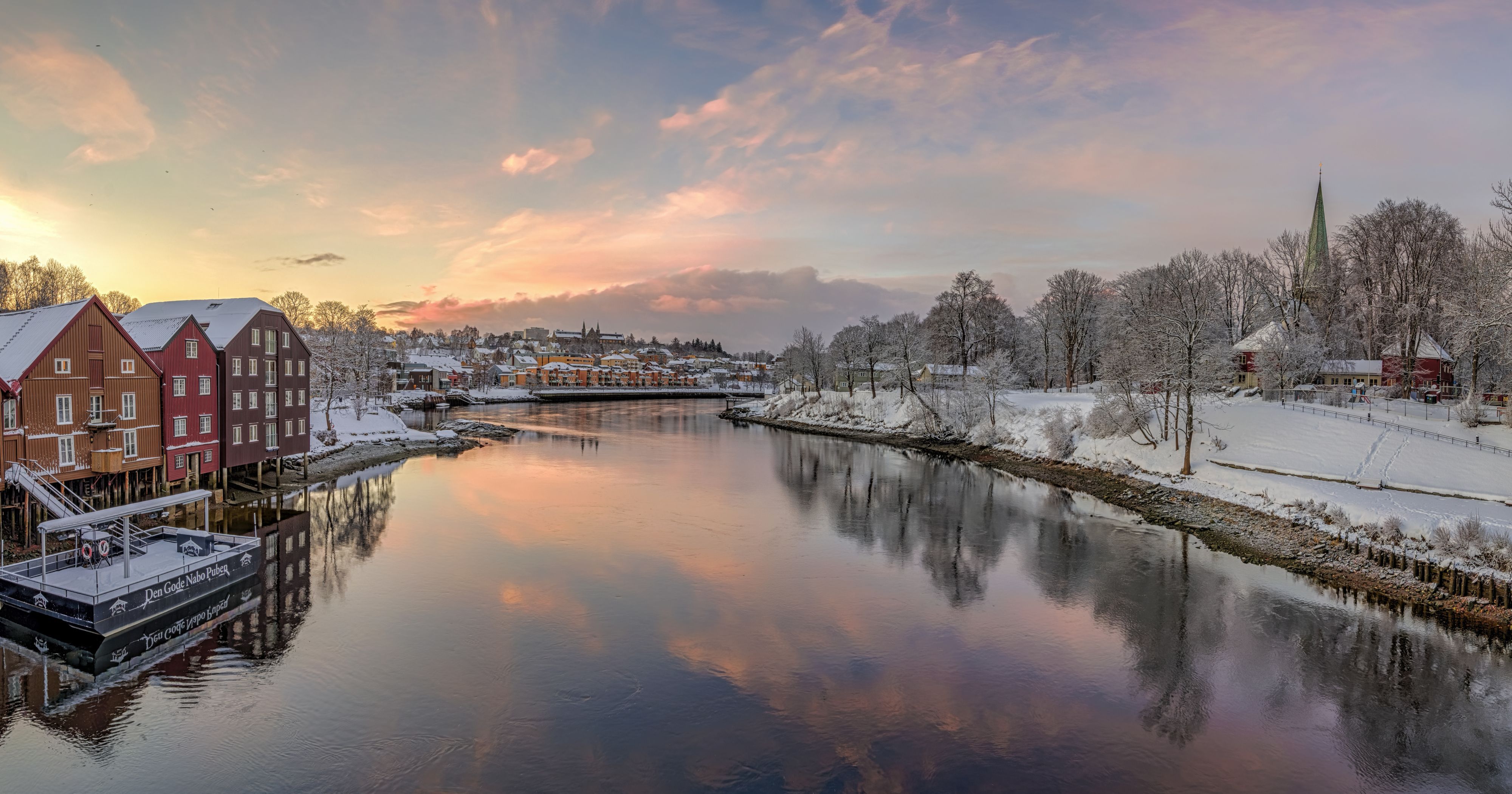 Snødekte bryggehus og Nidarosdomens spir speiler seg i Nidelva under en rosa vinter-solnedgang i Trondheim, Norge