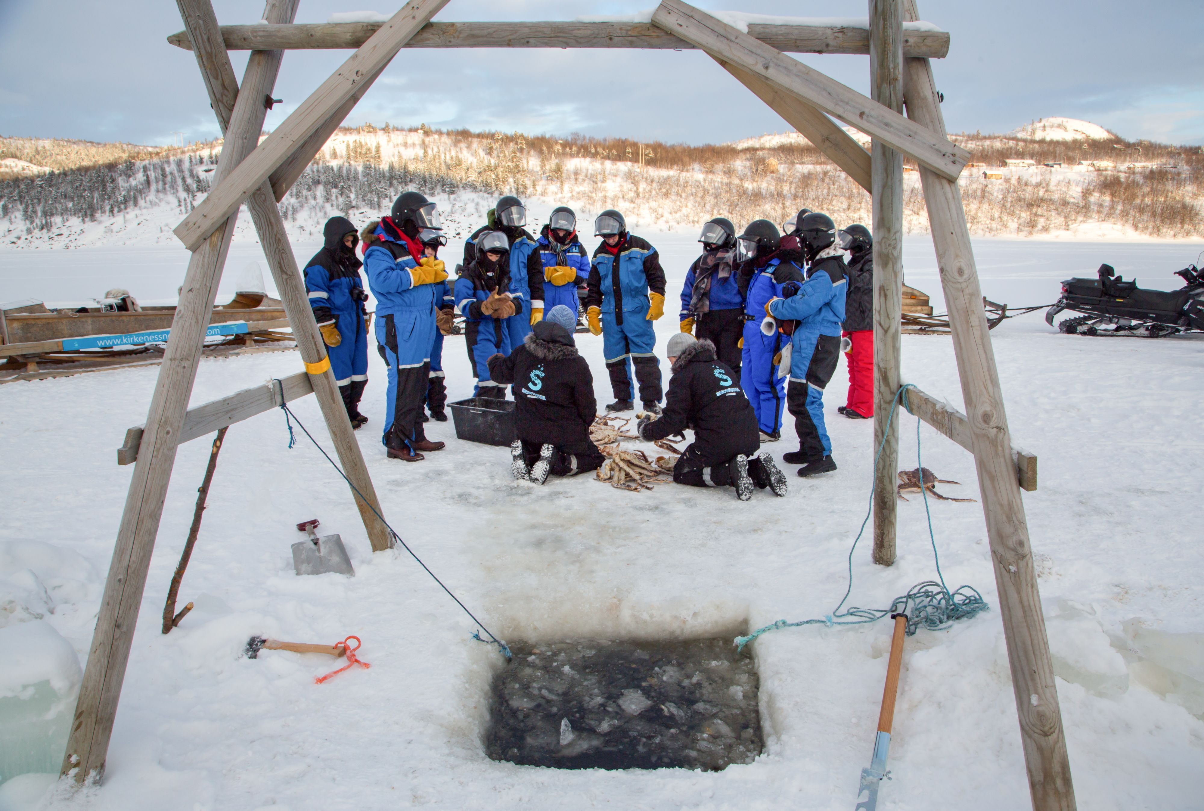 Gjester samles rundt et isfiskehull under en vinterlig kongekrabbsafari på en frossen fjord i Kirkenes, Norge.
