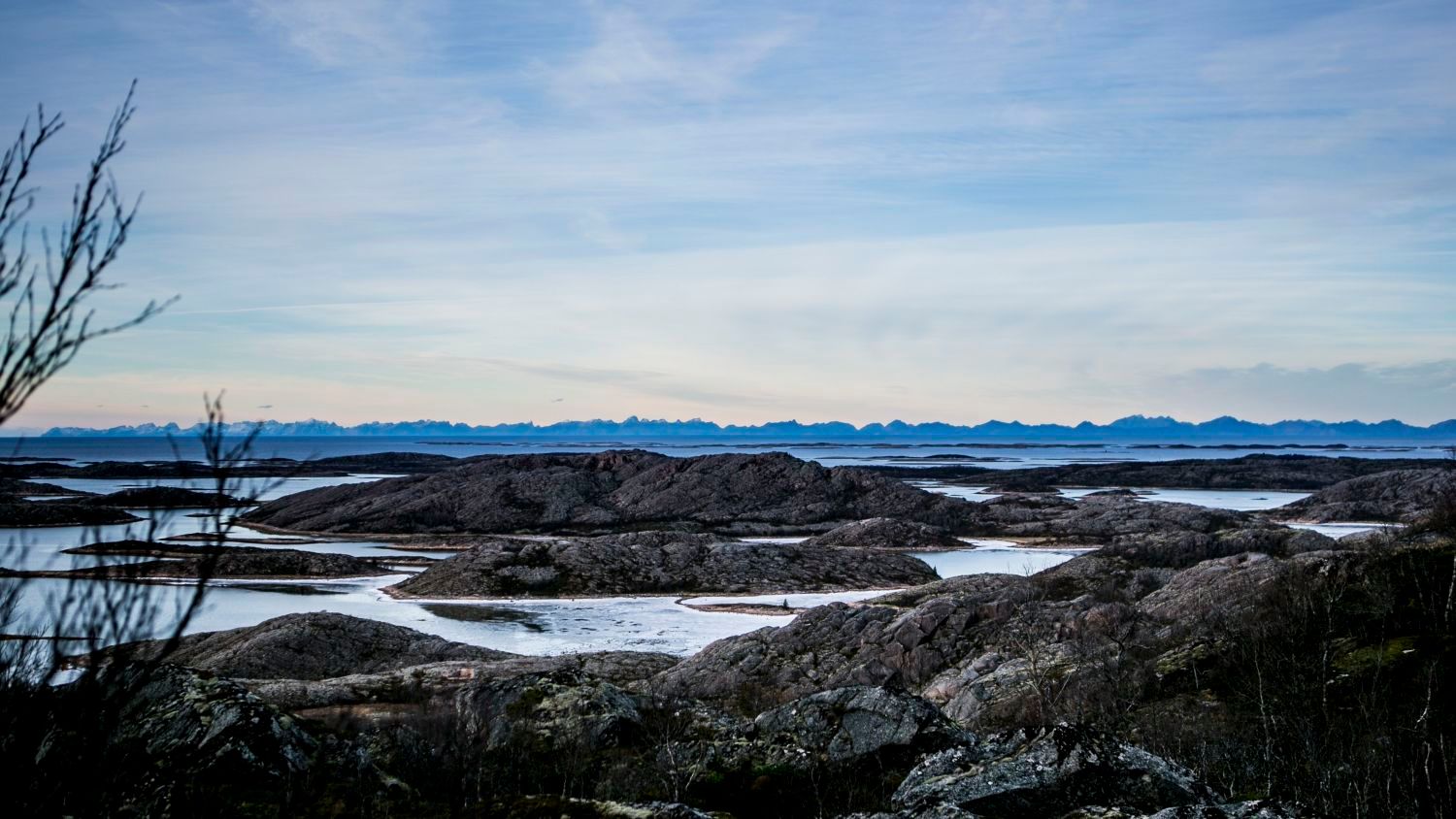 Felsige Schären und kleine Inseln entlang des Vestfjords mit der fernen Lofoten-Gebirgskette unter einem ruhigen blauen Himmel in Nordnorwegen.