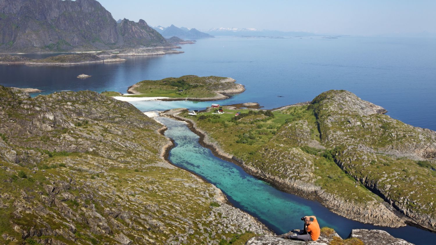 Türkise Küstenlagune und weißer Sandstrand auf den Lofoten in Norwegen, umgeben von zerklüfteten Bergen, felsigen Inseln und kristallklarem arktischem Wasser.
