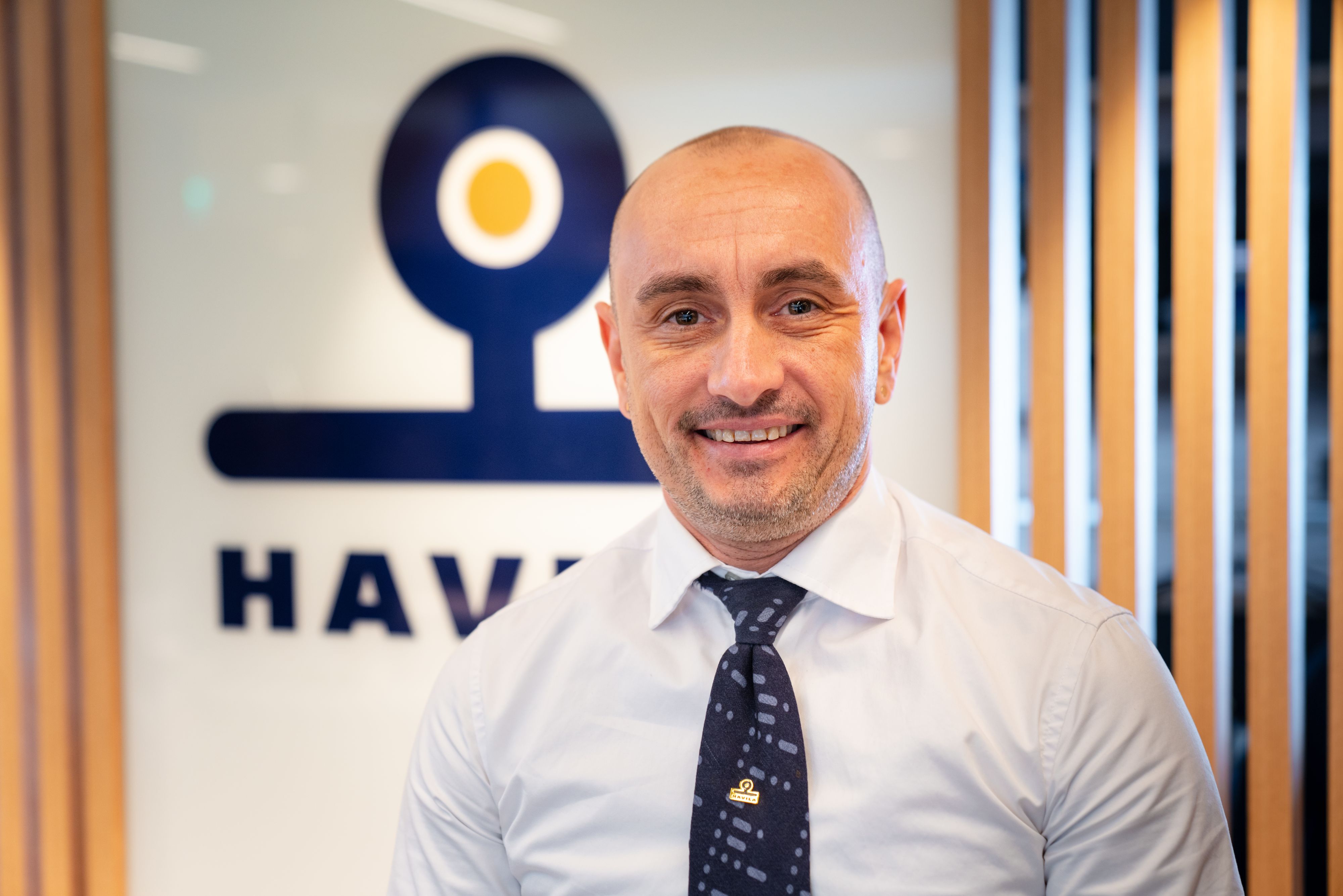 Smiling Havila Voyages crew member in uniform with the Havila logo in the background.