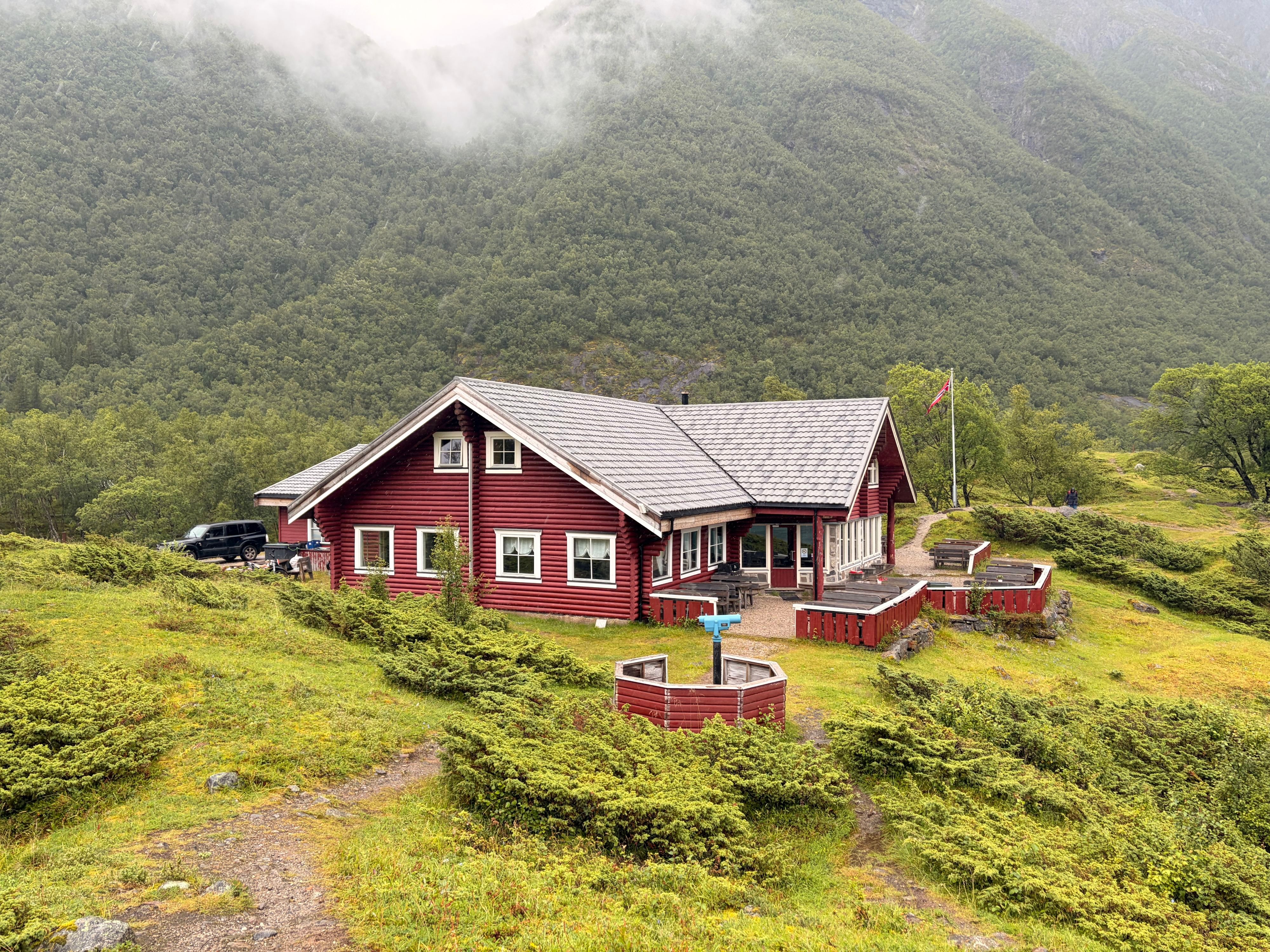 Rote Holzhütte mit norwegischer Flagge am Aussichtspunkt des Svartisen-Gletschers im Holandsfjord, Nebelschwaden ziehen über bewaldete Berge