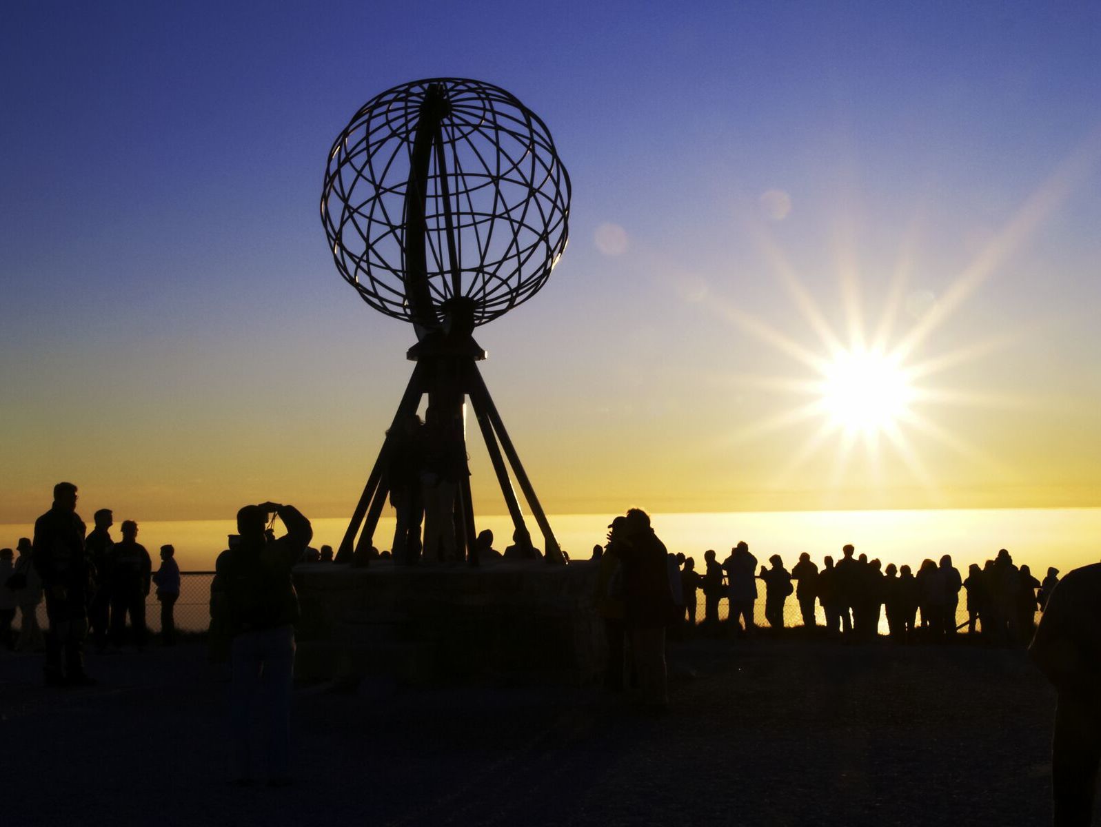 Besøkende samler seg rundt det ikoniske Nordkapp-globusmonumentet under midnattssolen, silhuettert mot en gyllen arktisk horisont på Europas nordligste punkt.