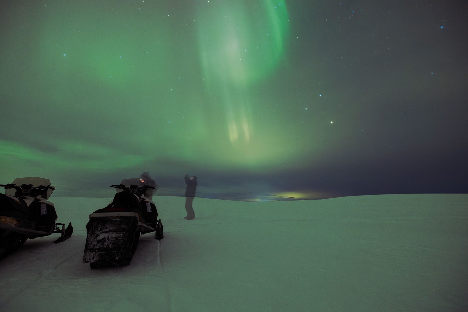 Tourist taking photo of northern lights next to two snow mobiles