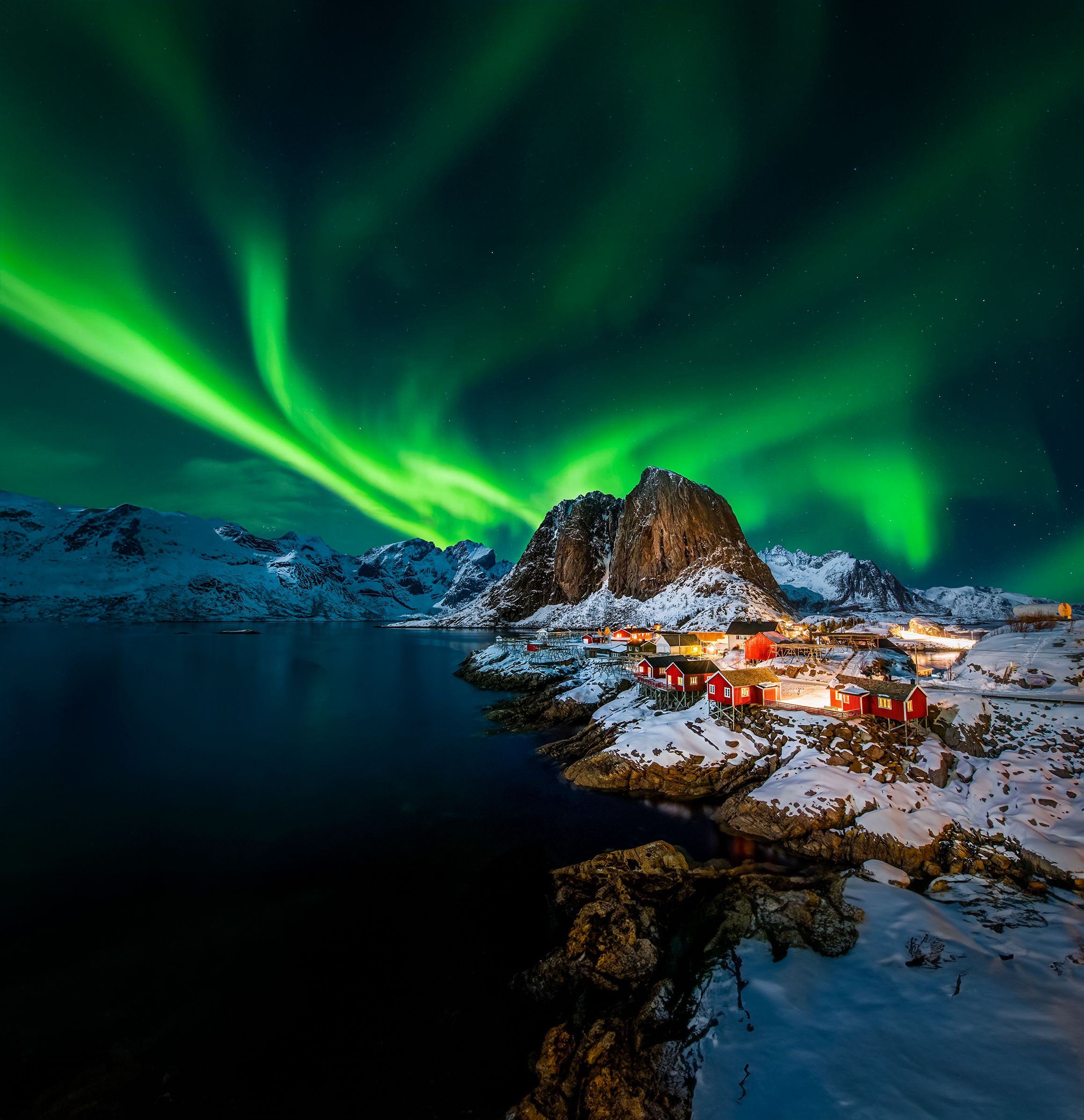 Nordlys over røde rorbuer og snødekte fjelltopper ved Reine i Lofoten