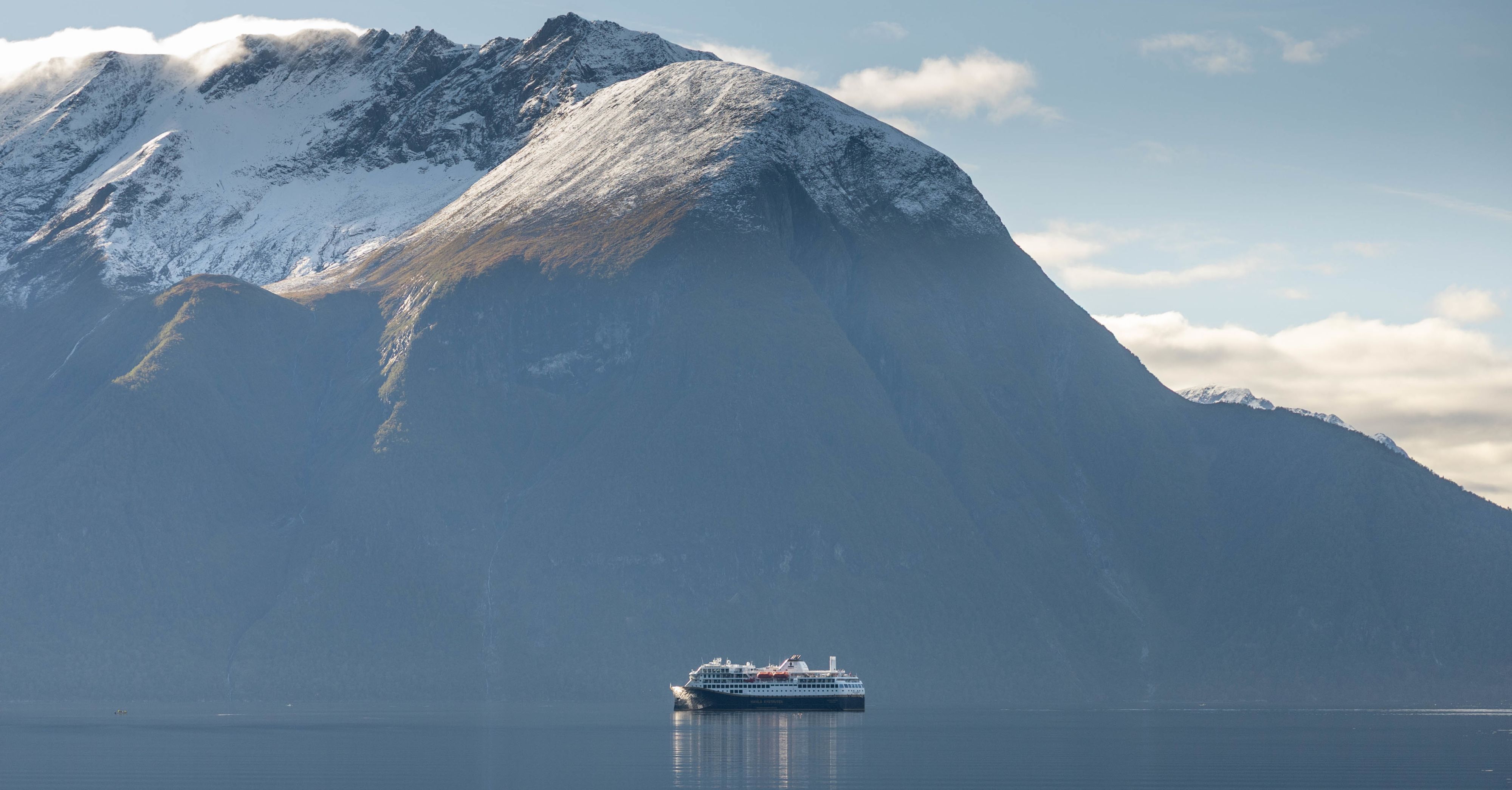 Ein Schiff von Havila Voyages fährt entlang der schneebedeckten Berge an Norwegens Küste und symbolisiert die Auszeichnung des Unternehmens bei den Grand Travel Awards 2024 als beste Fährreederei mit Verbindungen in oder nach Norwegen.