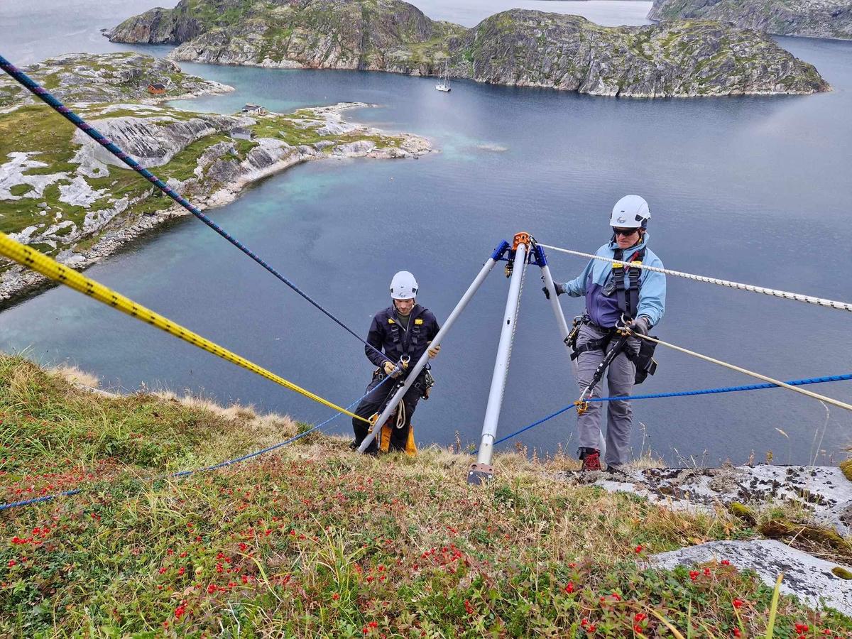 Two participants wearing helmets and climbing gear on an Arctic rappelling route above a rugged coastal landscape.