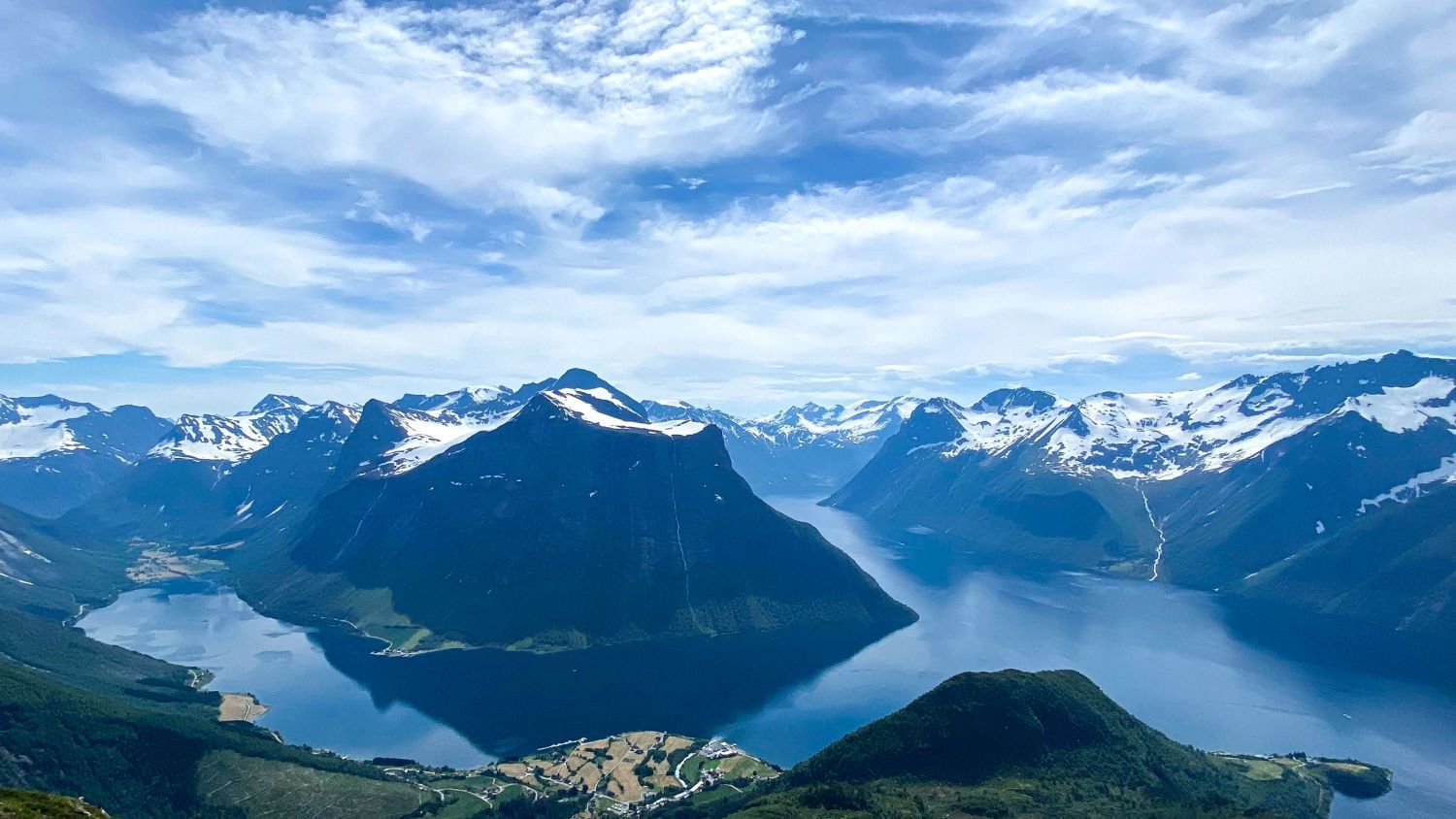 Panoramablick auf den Hjørundfjord, umgeben von den schneebedeckten Sunnmøre-Alpen, mit tiefblauem Fjordwasser, dramatischen Berggipfeln und unberührten Landschaften in Westnorwegen.