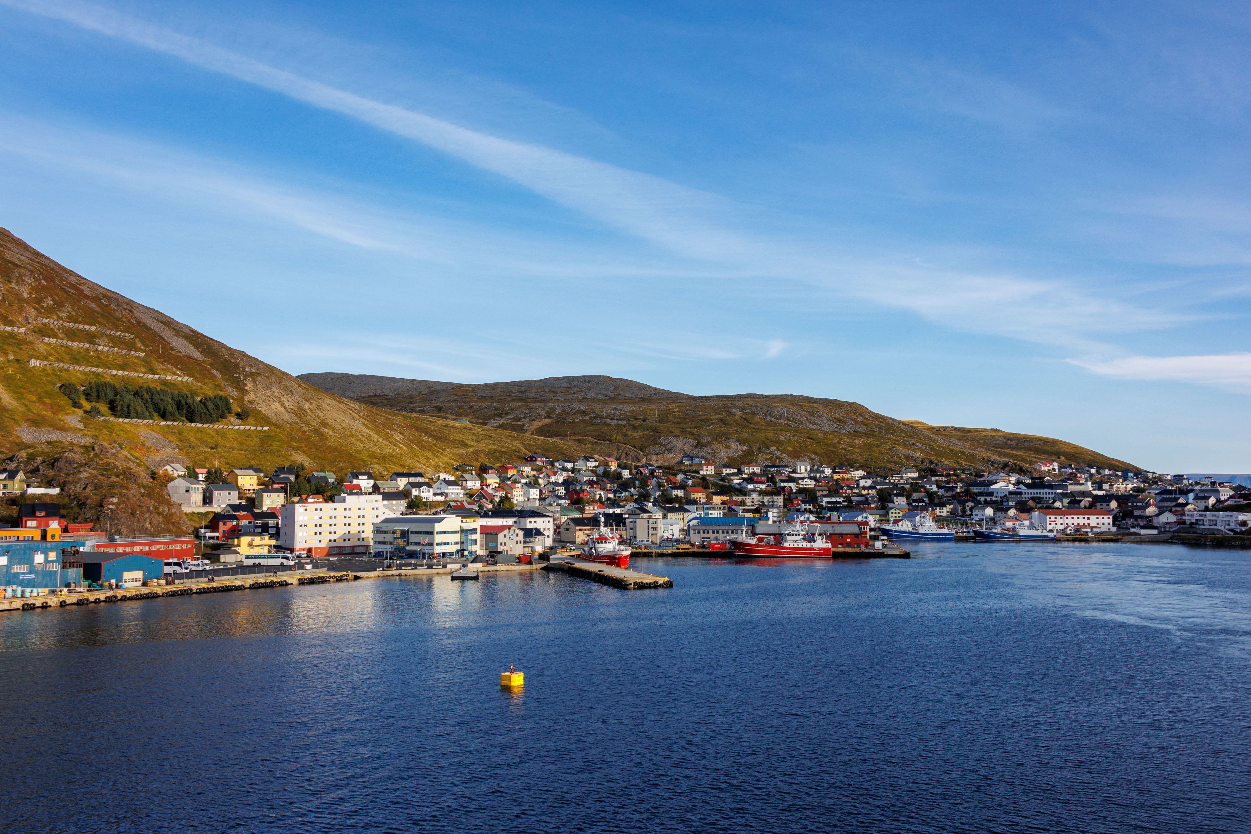 Panoramablick auf den Hafen von Honningsvåg mit bunten Gebäuden an der Uferpromenade, Fischerbooten und sanft geschwungenen arktischen Hügeln unter einem strahlend blauen Himmel – das Tor zum Nordkap.