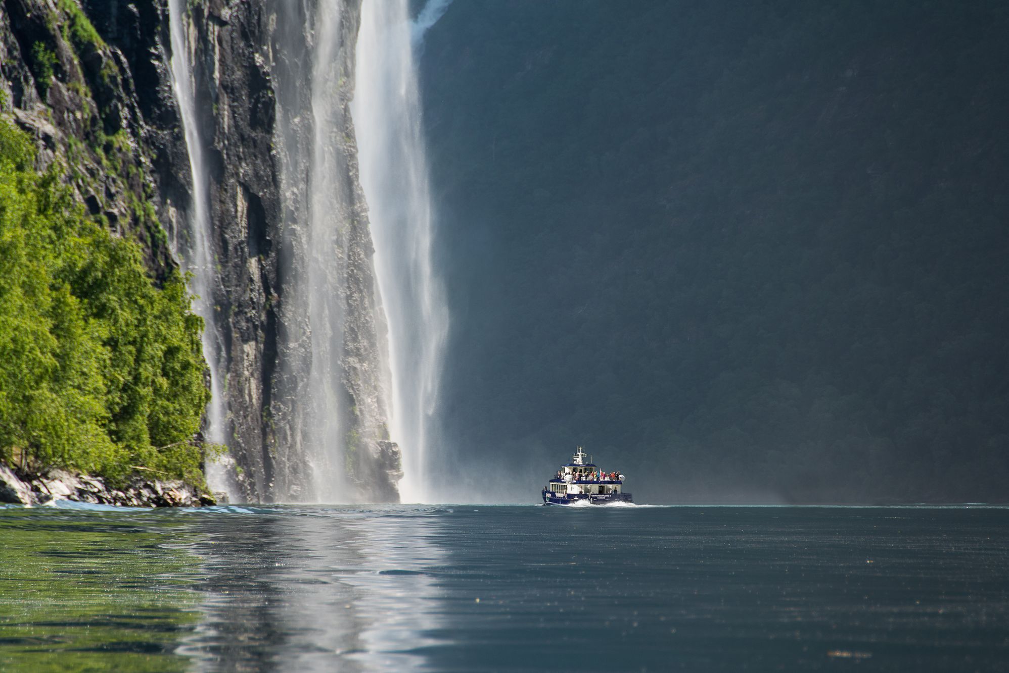 Sightseeing boat cruising beneath towering waterfalls in Geirangerfjord, surrounded by steep cliffs and dramatic Norwegian fjord scenery.