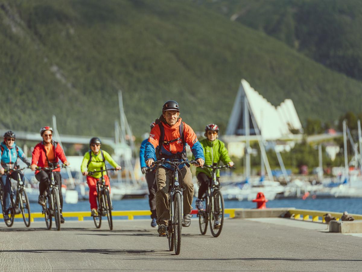 En gruppe syklister som kjører elsykler langs havnepromenaden i Tromsø, med fjell i bakgrunnen.