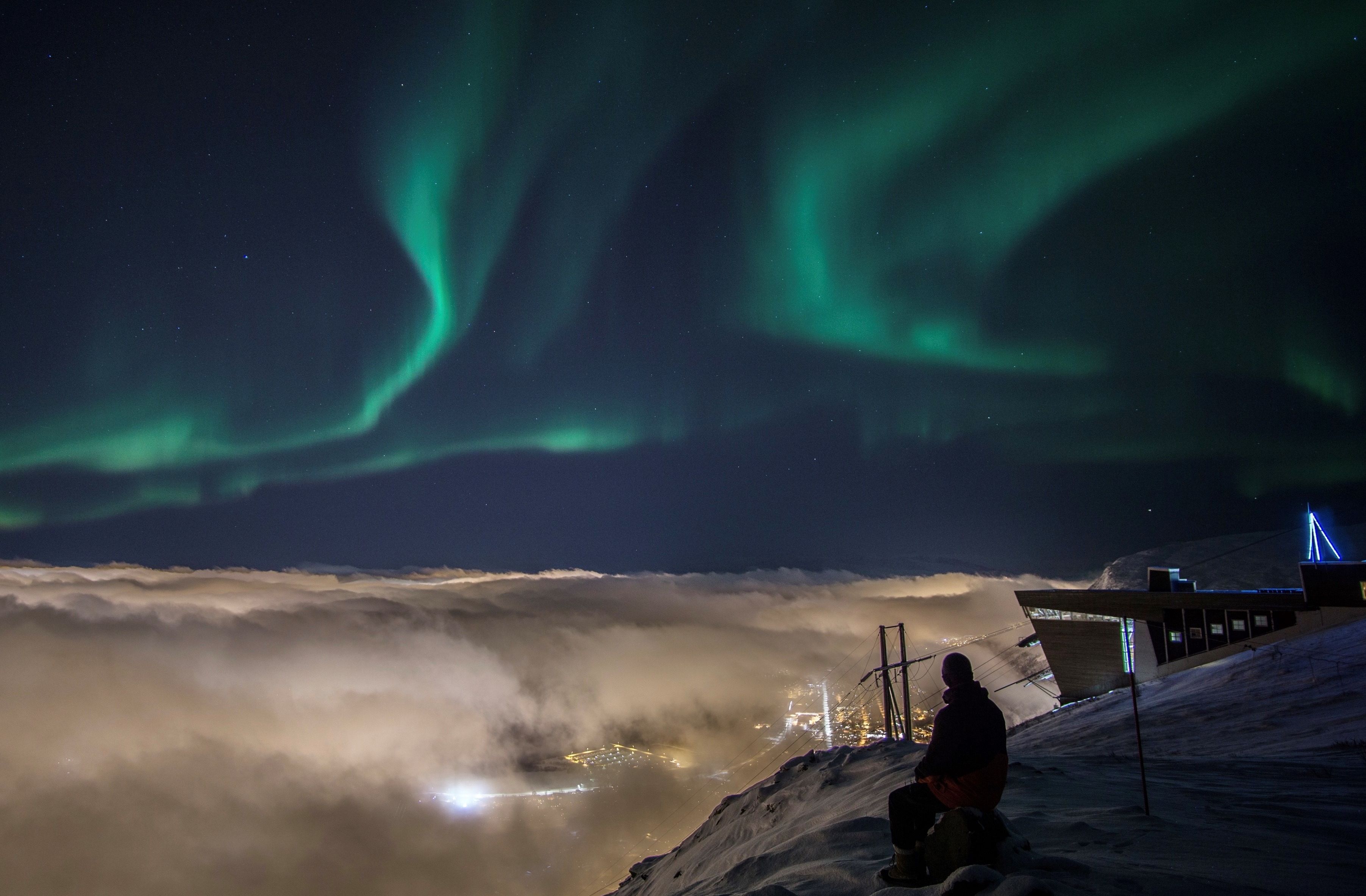 Person som ser på nordlyset fra Fjellheisen over Tromsø, med skyer som dekker byen nedenfor