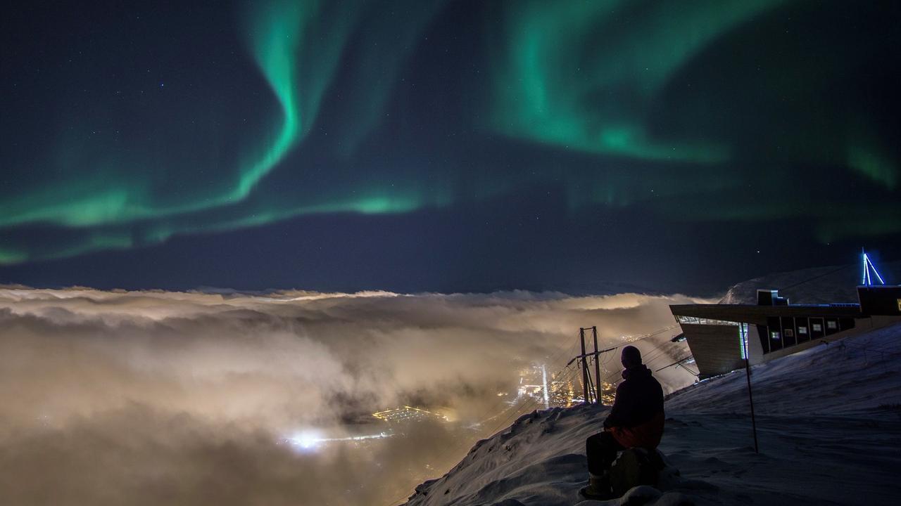 Person som betraktar norrskenet från Fjellheisen ovanför Tromsø, medan molnen täcker staden nedanför