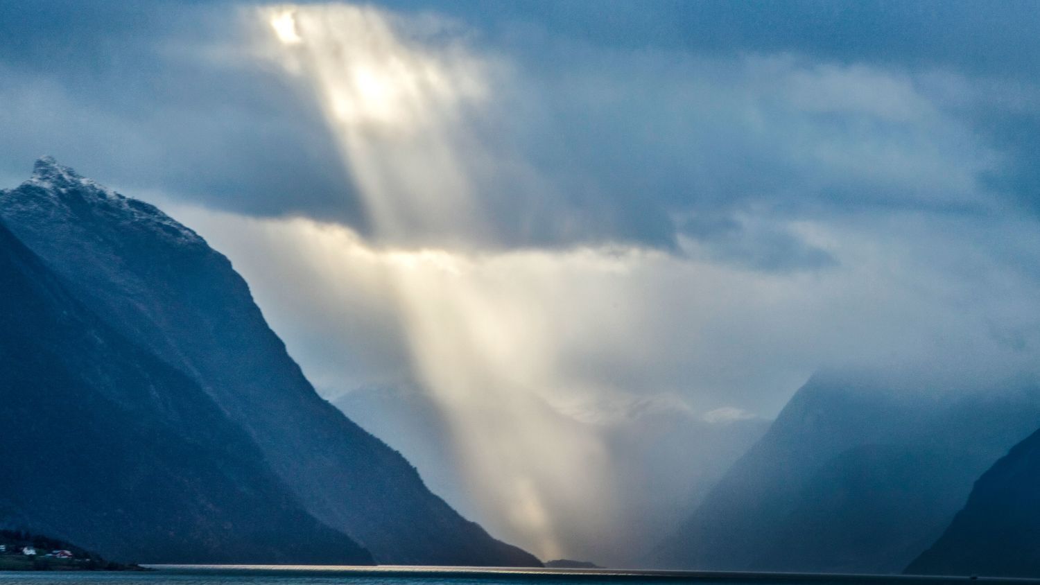 Sonnenstrahlen, die durch dramatische Sturmwolken über dem Hjørundfjord brechen und den majestätischen Fjord- und Gebirgslandschaft im Westen Norwegens erleuchten.