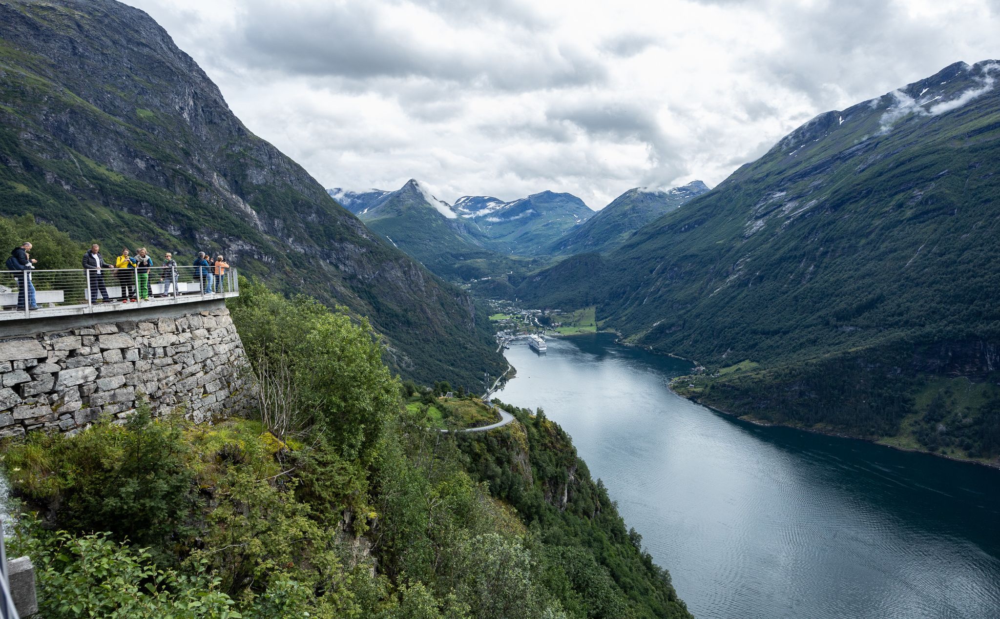 Besökare beundrar den panoramiska utsikten från utsiktsplatsen Flydalsjuvet med vy över Geirangerfjorden, de branta bergen, fjorden och byn Geiranger nedanför.
