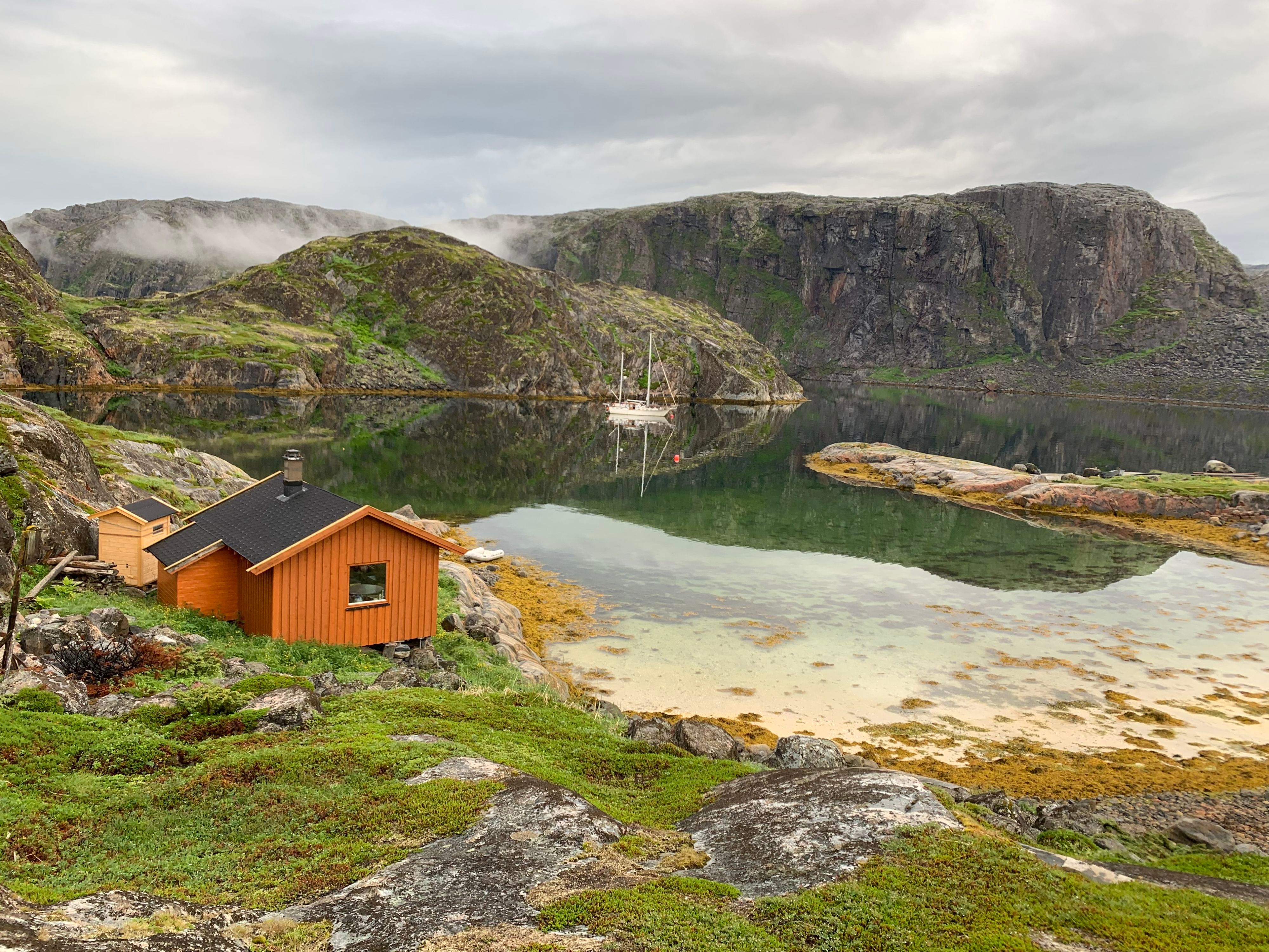 Peaceful Arctic fjord with a traditional cabin, crystal-clear water, and towering sea cliffs along the Arctic Rappelling Route in Northern Norway.