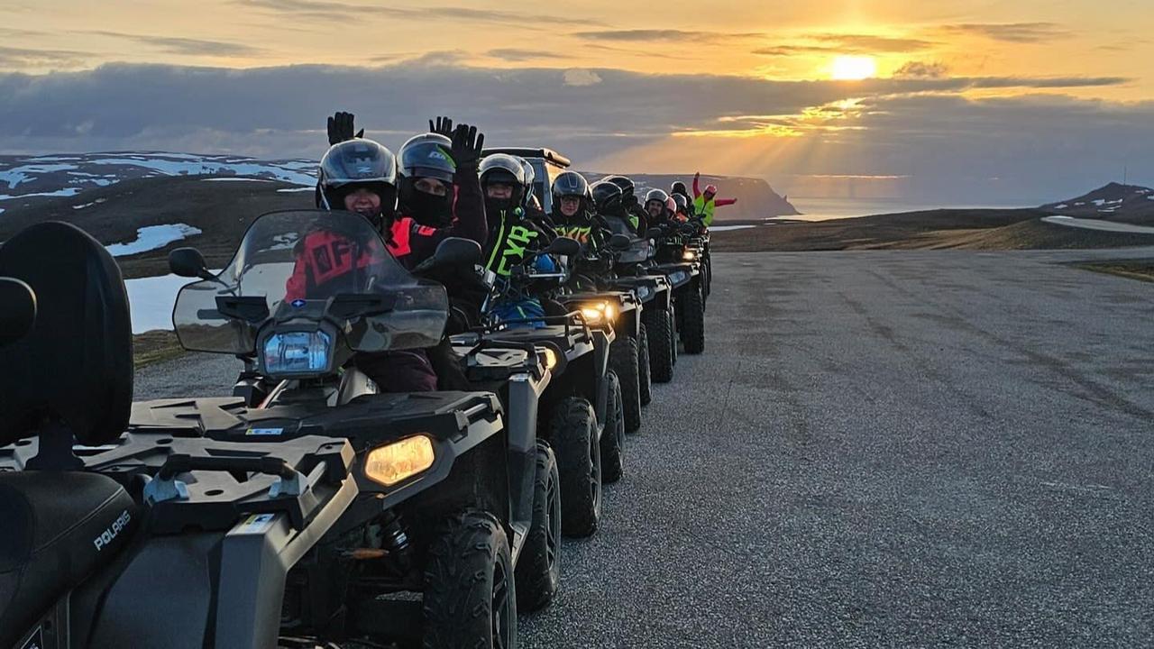 En grupp gäster på fyrhjulingar vinkar mot kameran under en guidad safari över den arktiska platån, med midnattssolen som lyser över landskapet på vägen mot Nordkap.