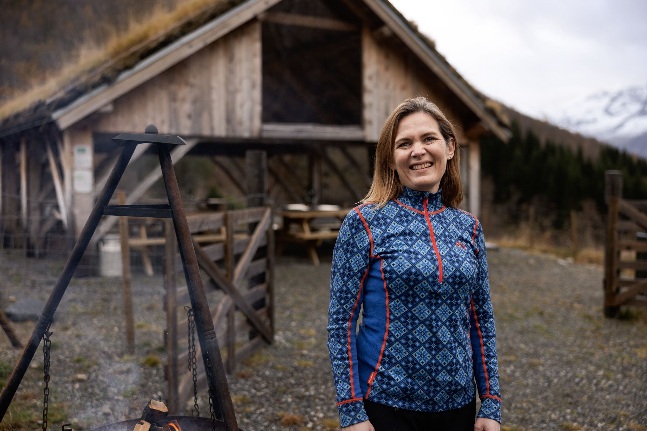 Lächelnde Frau in einem gemusterten Pullover am Lagerfeuer vor einer traditionellen Berghütte mit Grasdach auf einem Bergbauernhof im Hjørundfjord in Norwegen