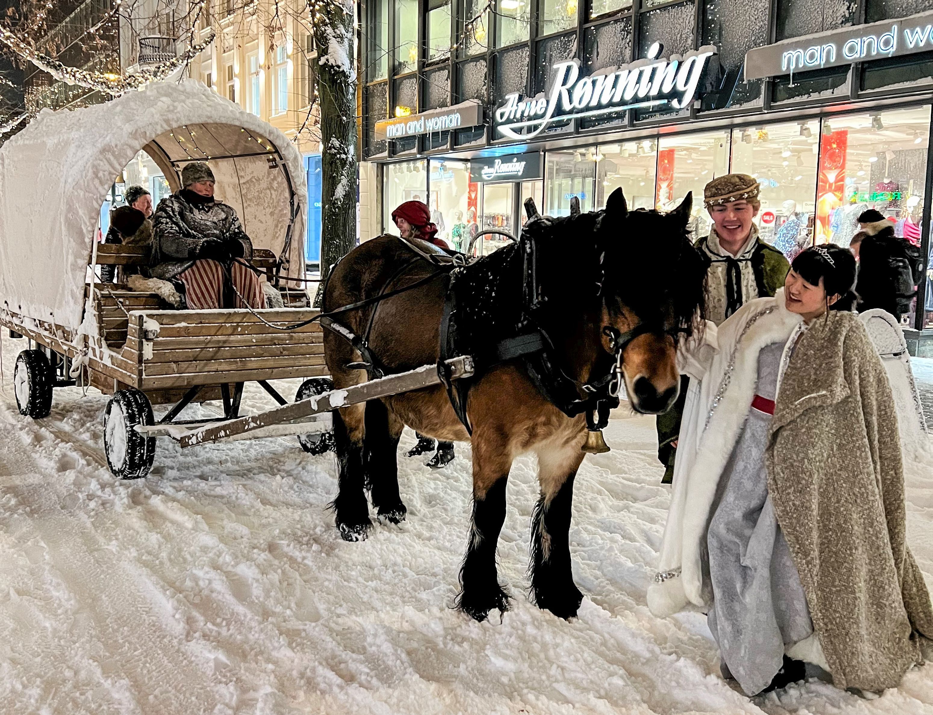 Horse-drawn carriage and costumed guides on a snowy street at the Trondheim Christmas market at night, Norway