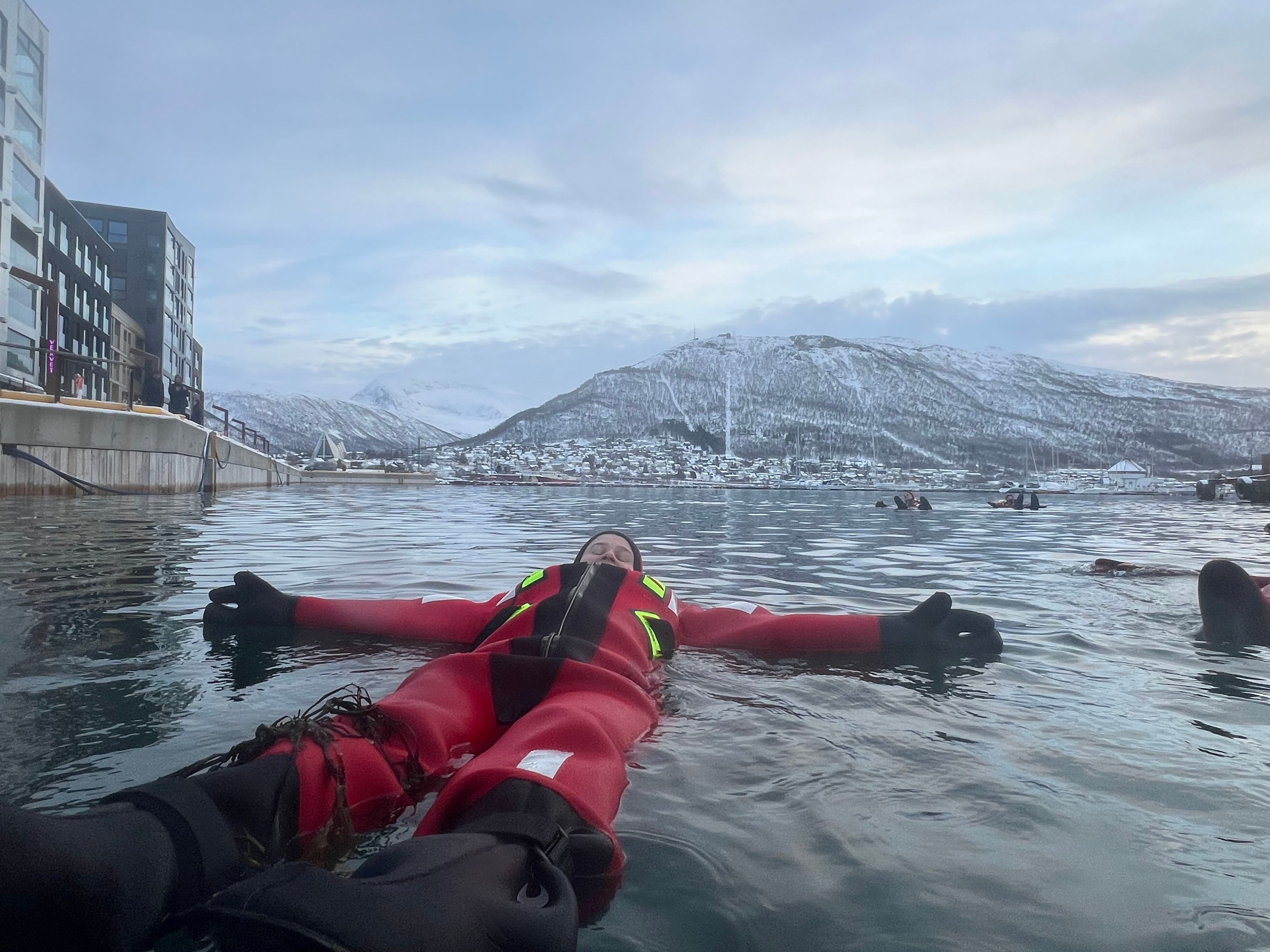 Arctic floating excursion in Tromsø, Norway, with participants relaxing in insulated suits on the calm waters beneath snow-covered mountains.