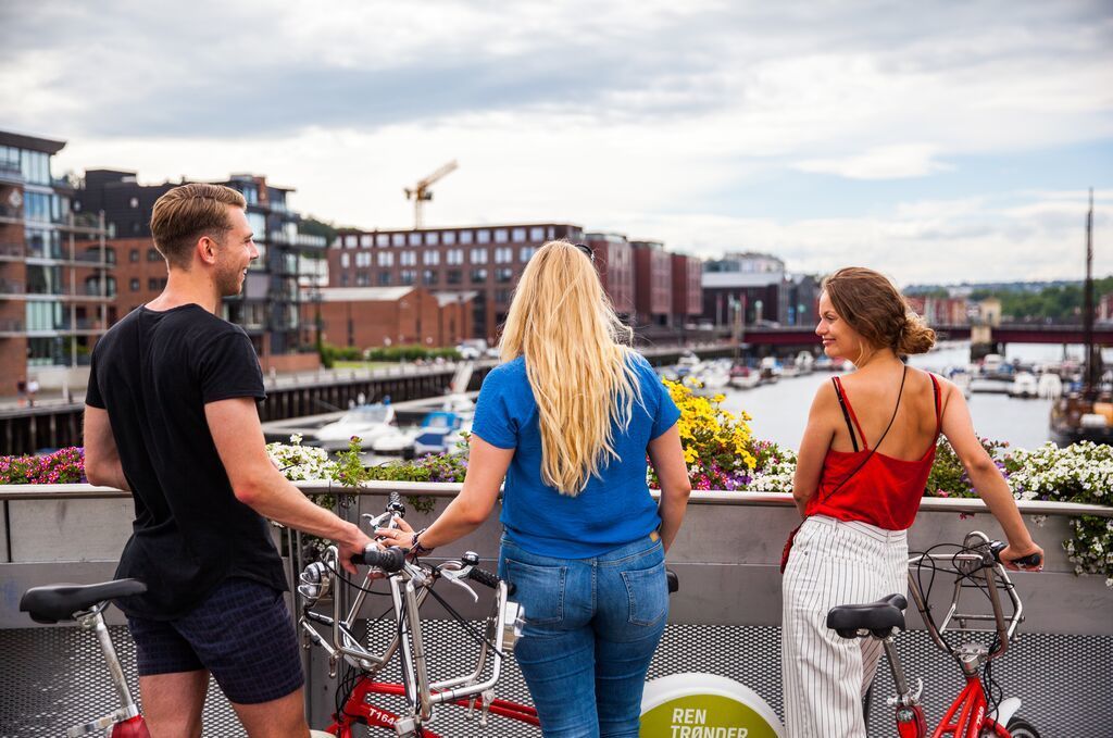 Tre venner med bysykler som nyter havnen og utsikten over vannet i Trondheim, Norge