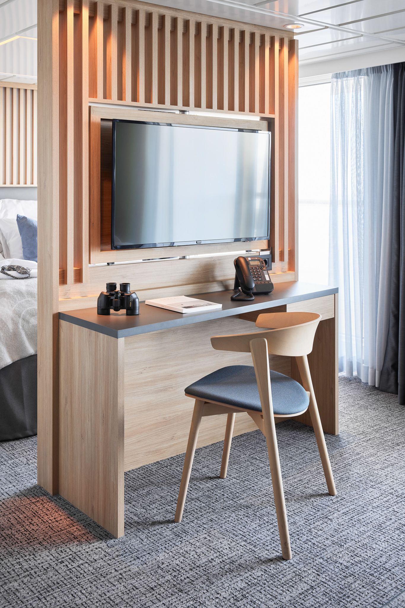 Desk with a chair beneath a swivel TV, featuring a telephone, binoculars, and a bright seating area beside floor-to-ceiling windows.