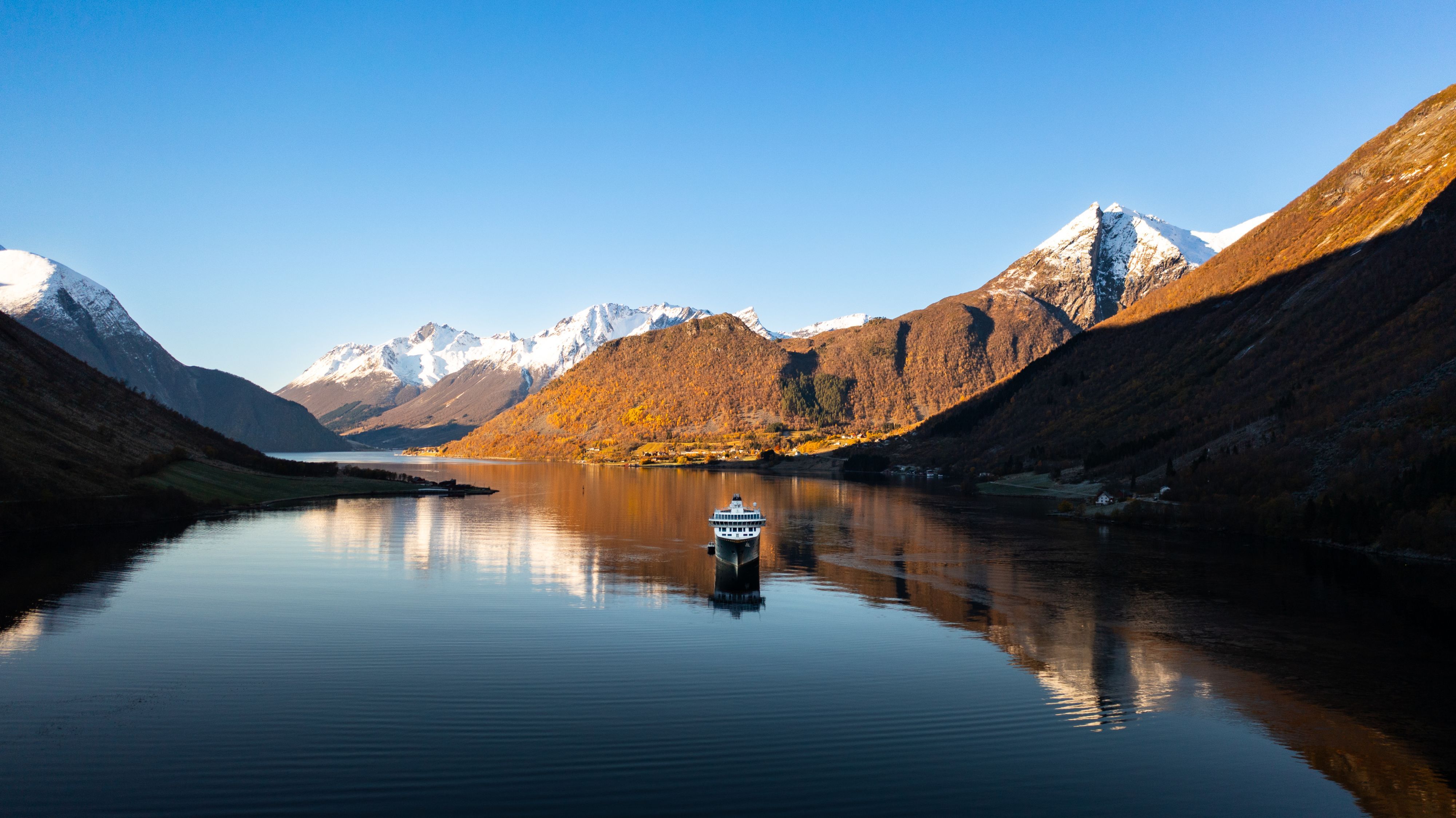 Havila-cruiseskip som seiler gjennom en rolig norsk fjord omgitt av høstfargede fjell og snødekte topper, Hjørundfjorden