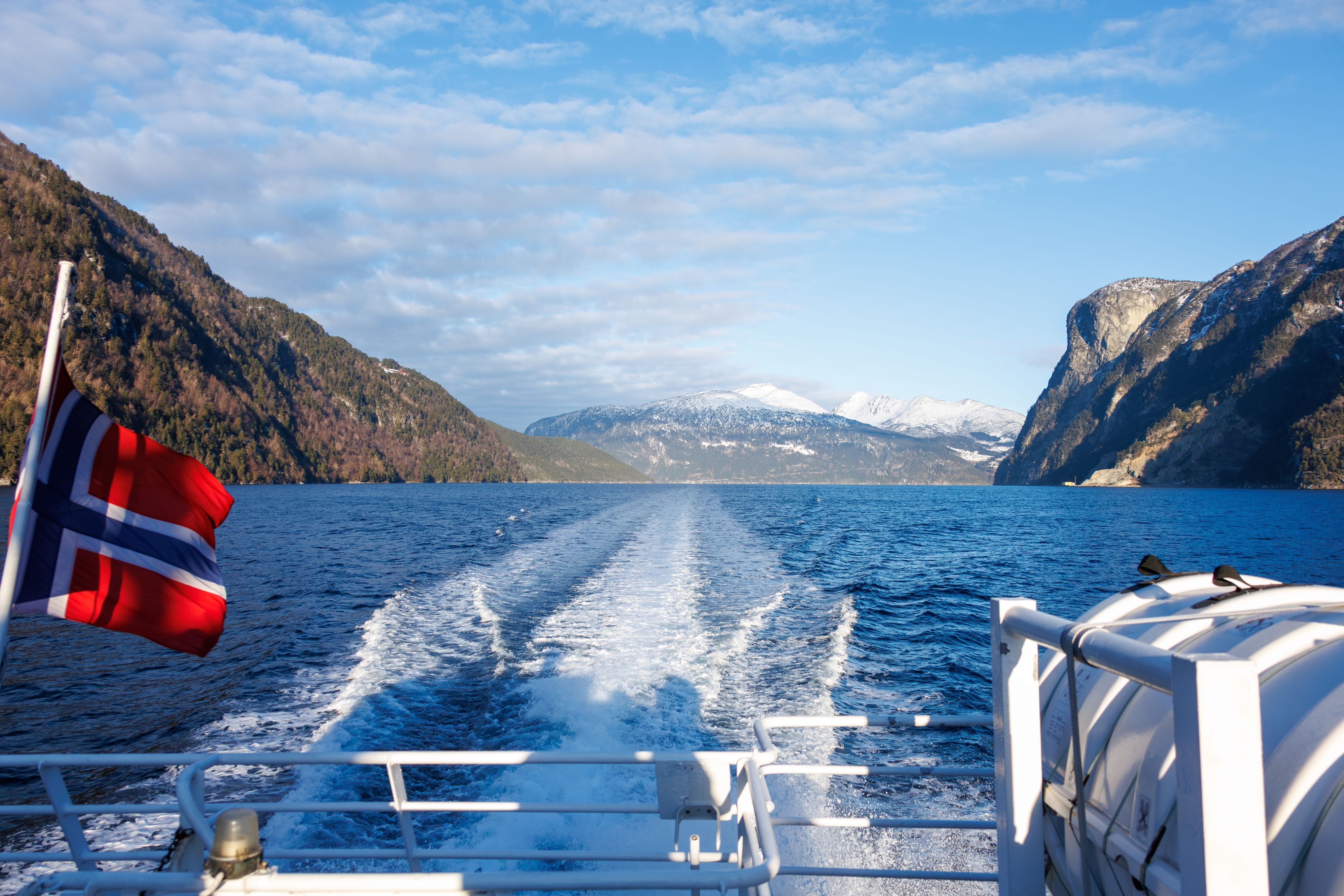 View from the stern of a fjord cruise boat with Norwegian flag and snowy mountains, Norway