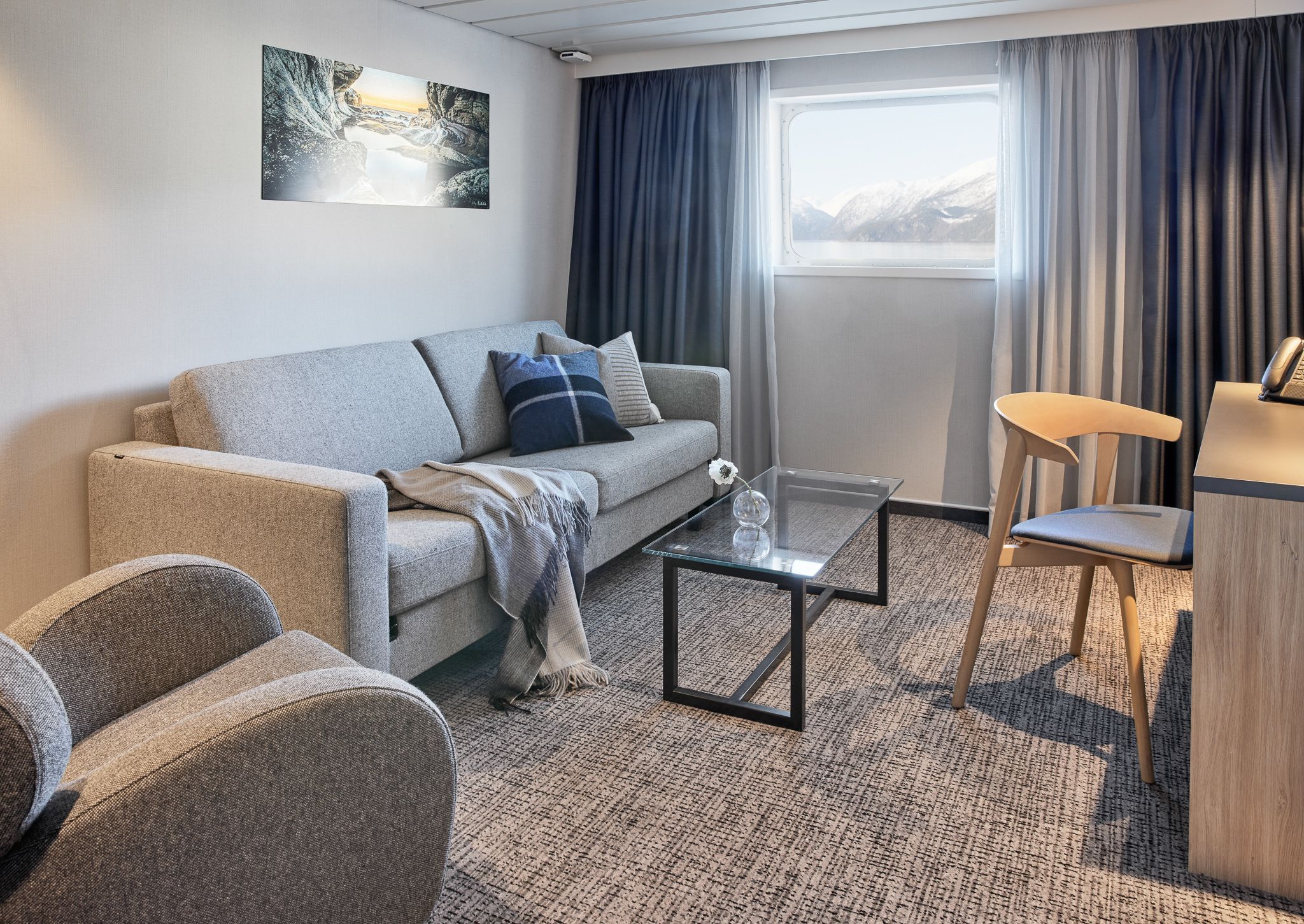 Seaview Juniorsuite med sittegruppe som inkluderer sovesofa, lenestol, salongbord, skrivebord og stort vindu med utsikt over sjøen og fjellene.