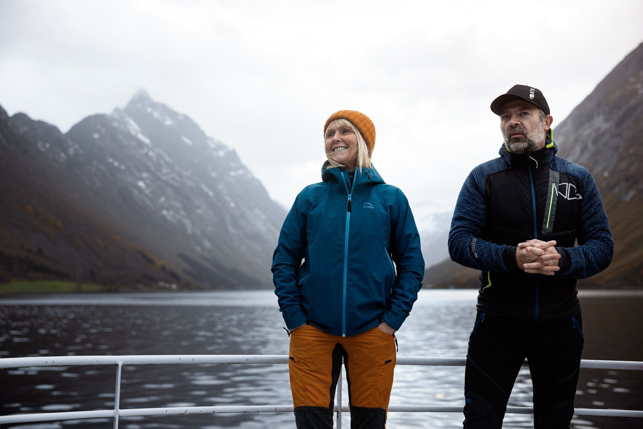 Zwei Personen in Outdoorjacken auf einem Schiffsdeck mit nebelverhangenen Bergen im Hintergrund während einer Bergwanderung im Hjørundfjord in Norwegen