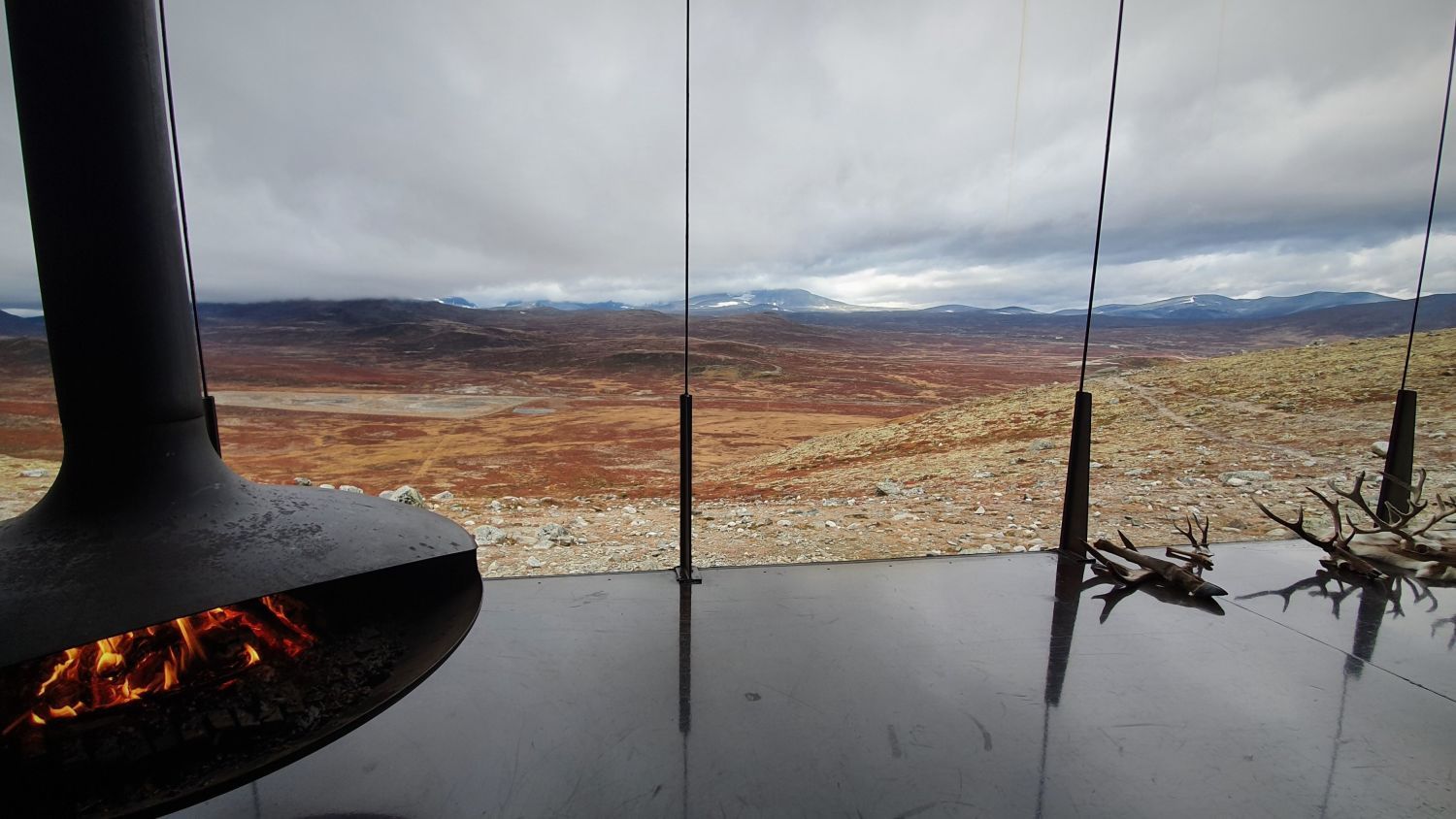 Panoramablick auf die norwegischen Berge von einer modernen Berghütte mit einem schwebenden Kamin, der auf die alpine Landschaft hinausblickt.