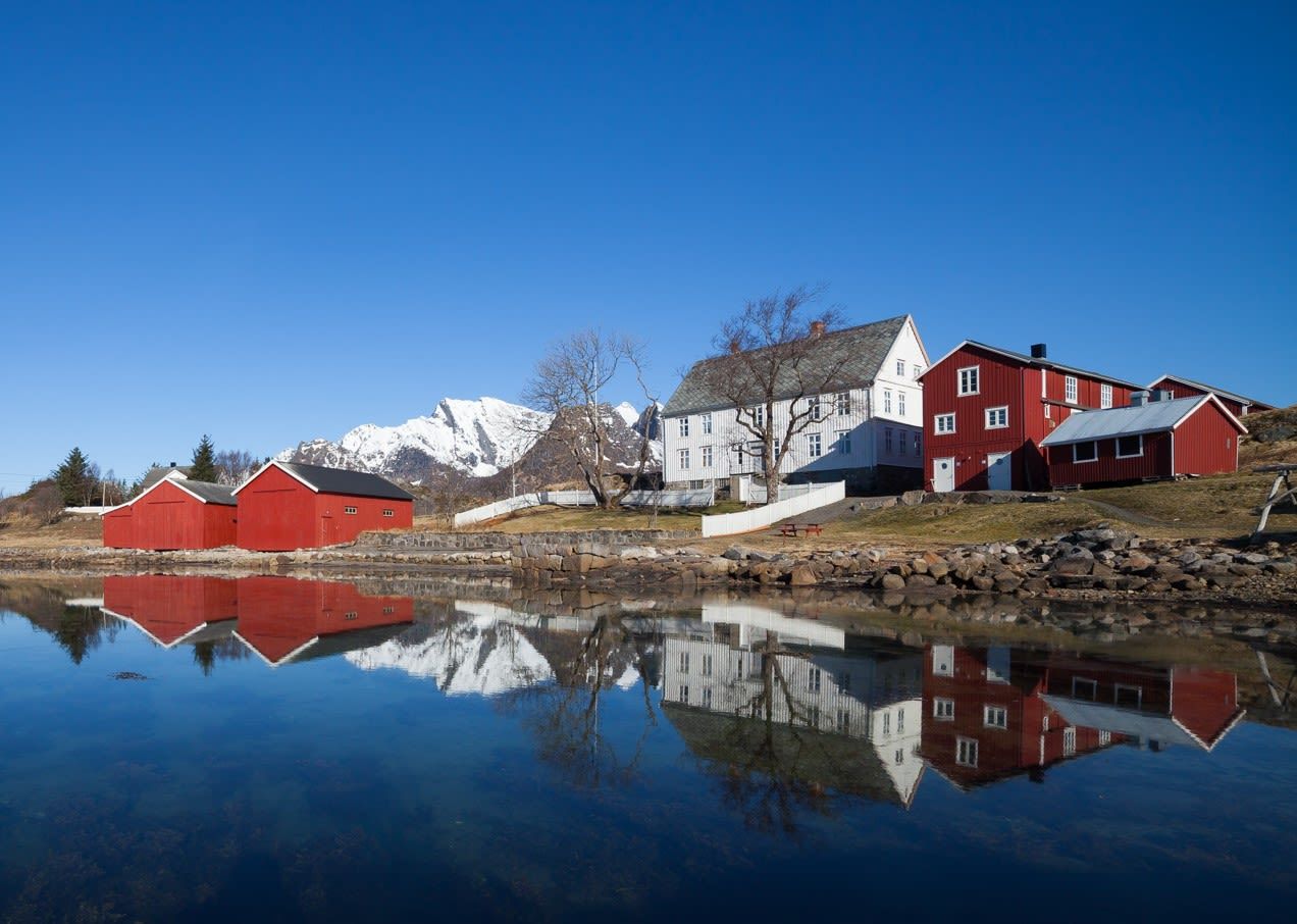 Traditional red and white houses reflected in calm fjord water with snowy mountains under a blue sky in Lofoten, Norway