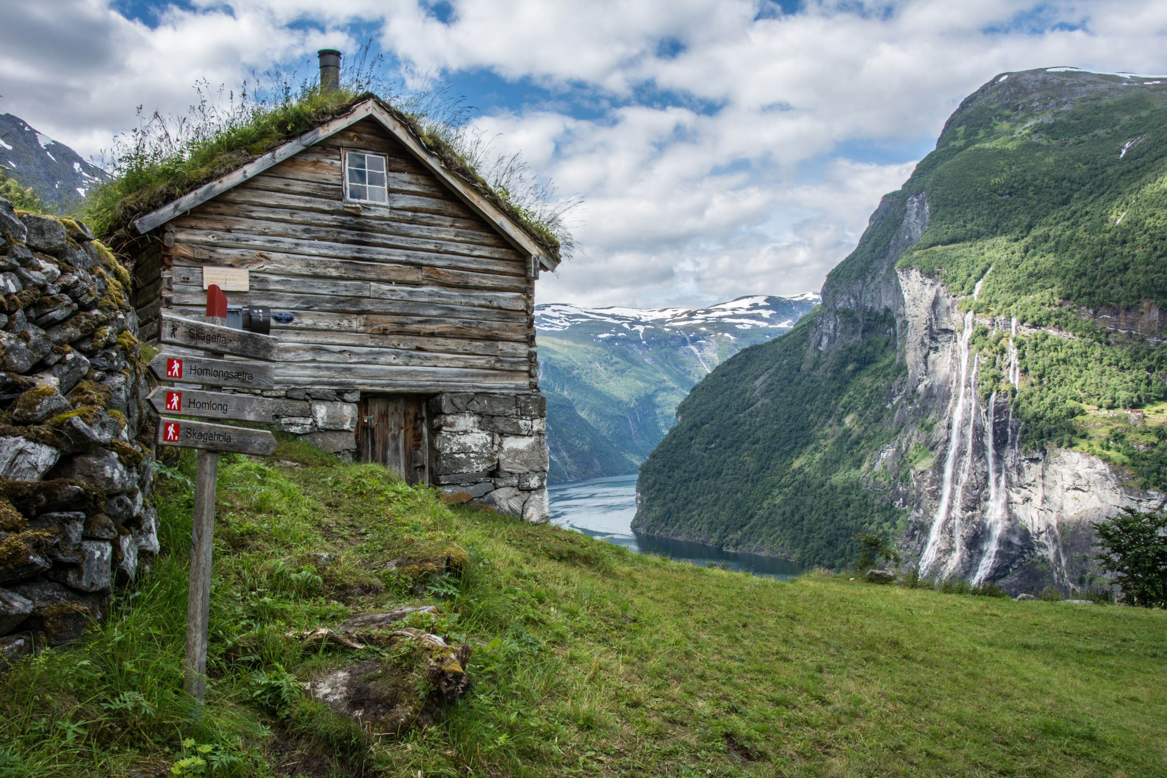 Historiske fjellgården Skageflå med utsikt over Geirangerfjorden, omgitt av stupbratte fjell, smaragdgrønt vann og fossen De sju søstrene i det fjerne
