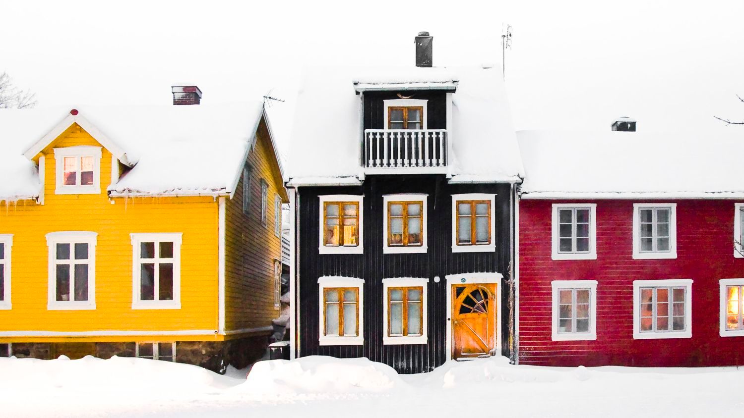 Bunte traditionelle Holzhäuser, gelb, schwarz und rot gestrichen, stehen Seite an Seite unter tief verschneiten Dächern und schaffen eine klassische winterliche Straßenszene in einem norwegischen Dorf.