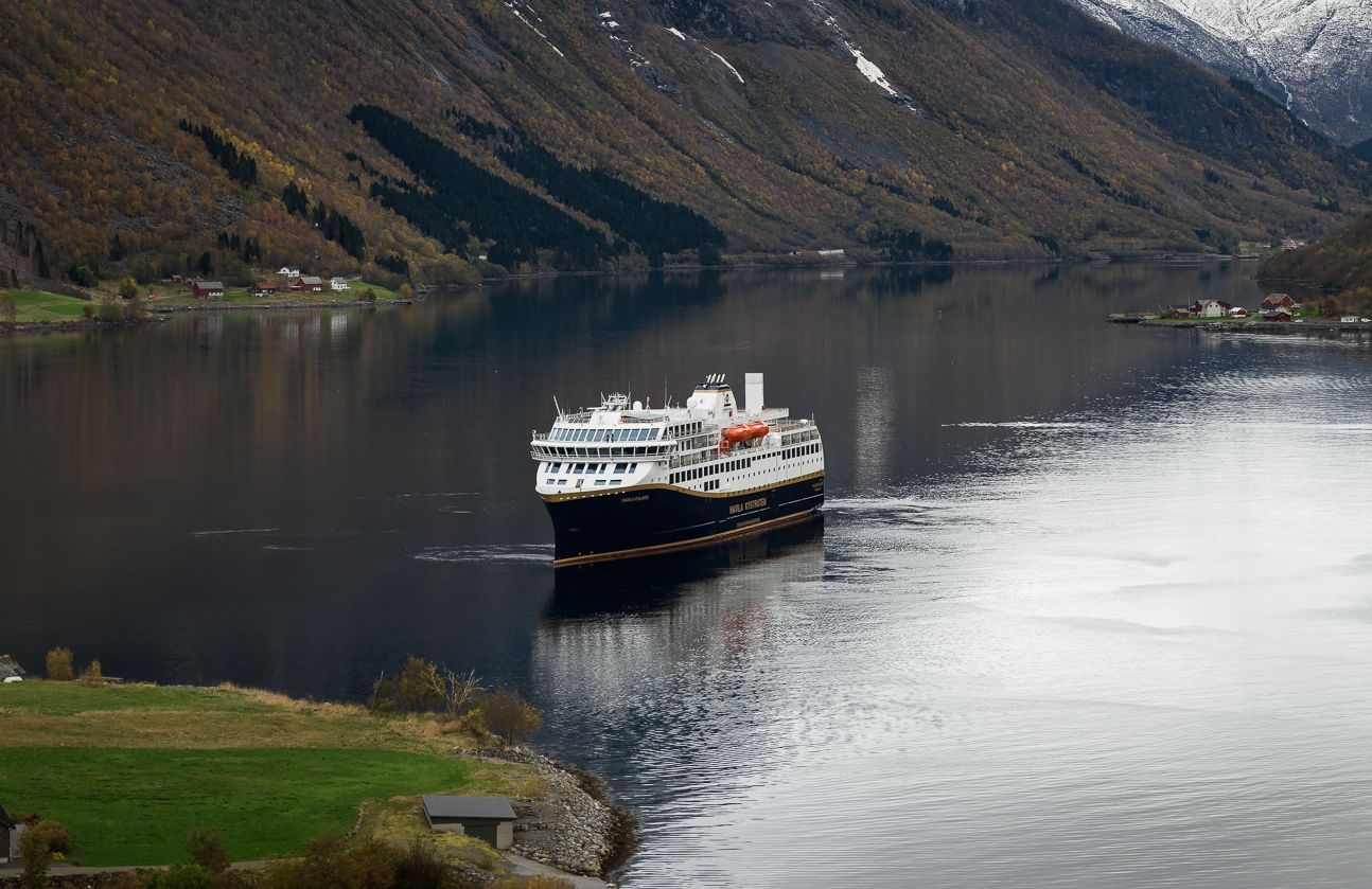 Havila-fartyg som seglar genom Hjørundfjords stilla vatten, omgivet av berg och fjordlandskap.