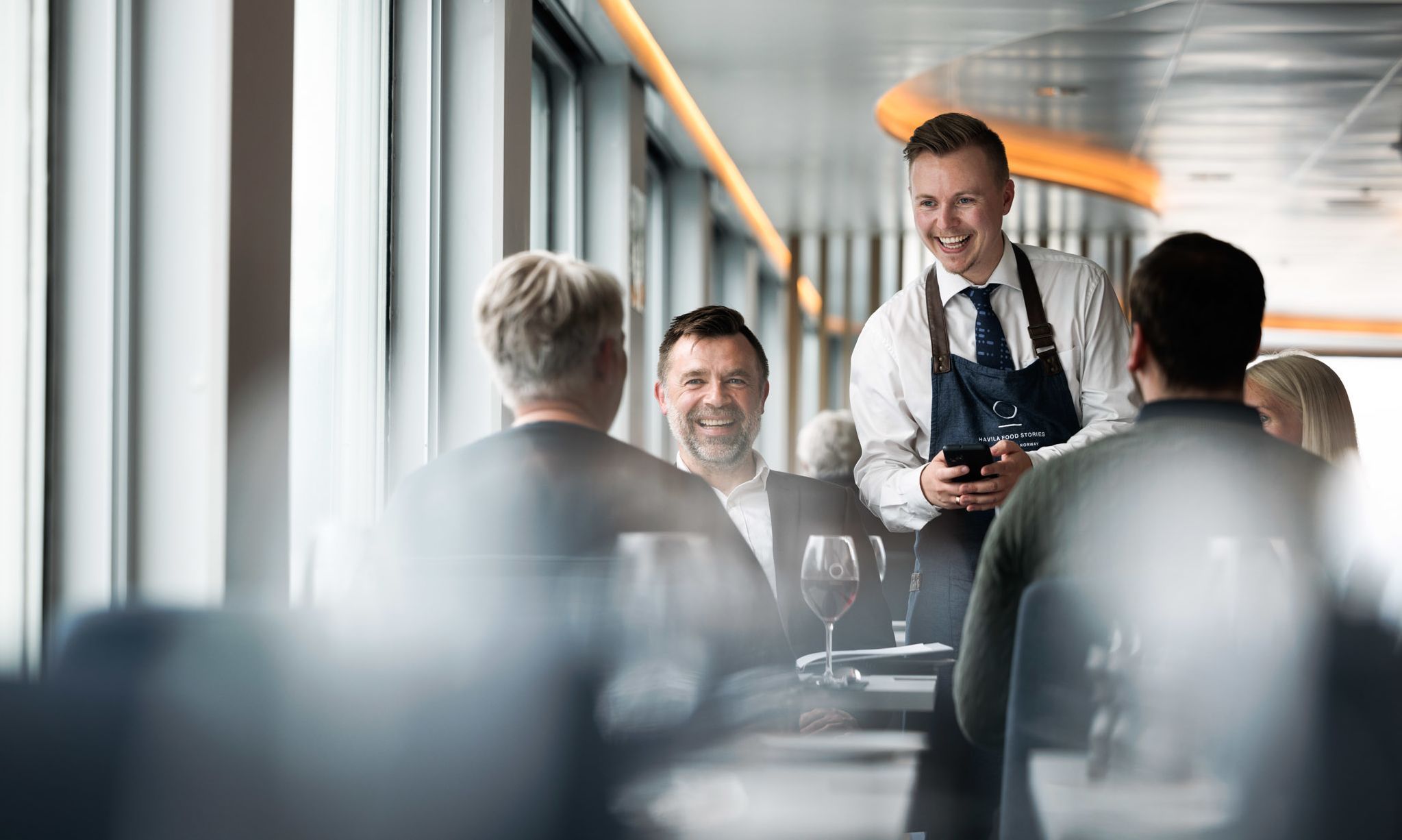 Guests enjoying attentive table service in the Havila Voyages restaurant with panoramic sea views.