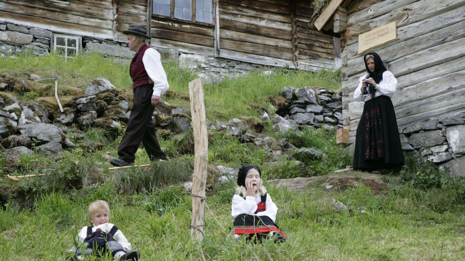 Familie in traditionellen norwegischen Volkstrachten vor historischen hölzernen Berghofgebäuden, die das ländliche Kulturerbe Norwegens zeigen.