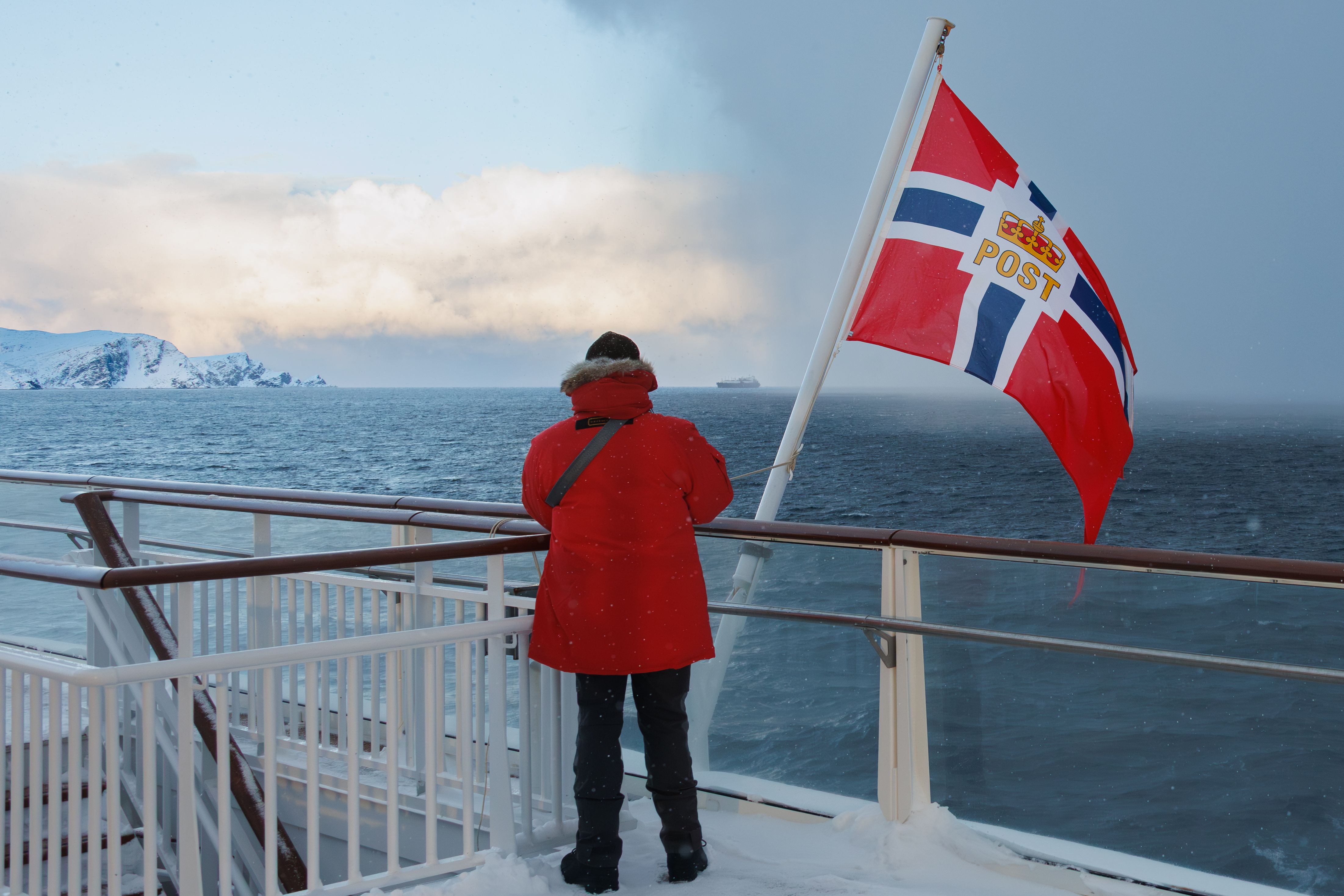 Gjest i rød parkas på et snødekt dekk ved siden av det norske postflagget, med blikket rettet mot snøkledde fjell