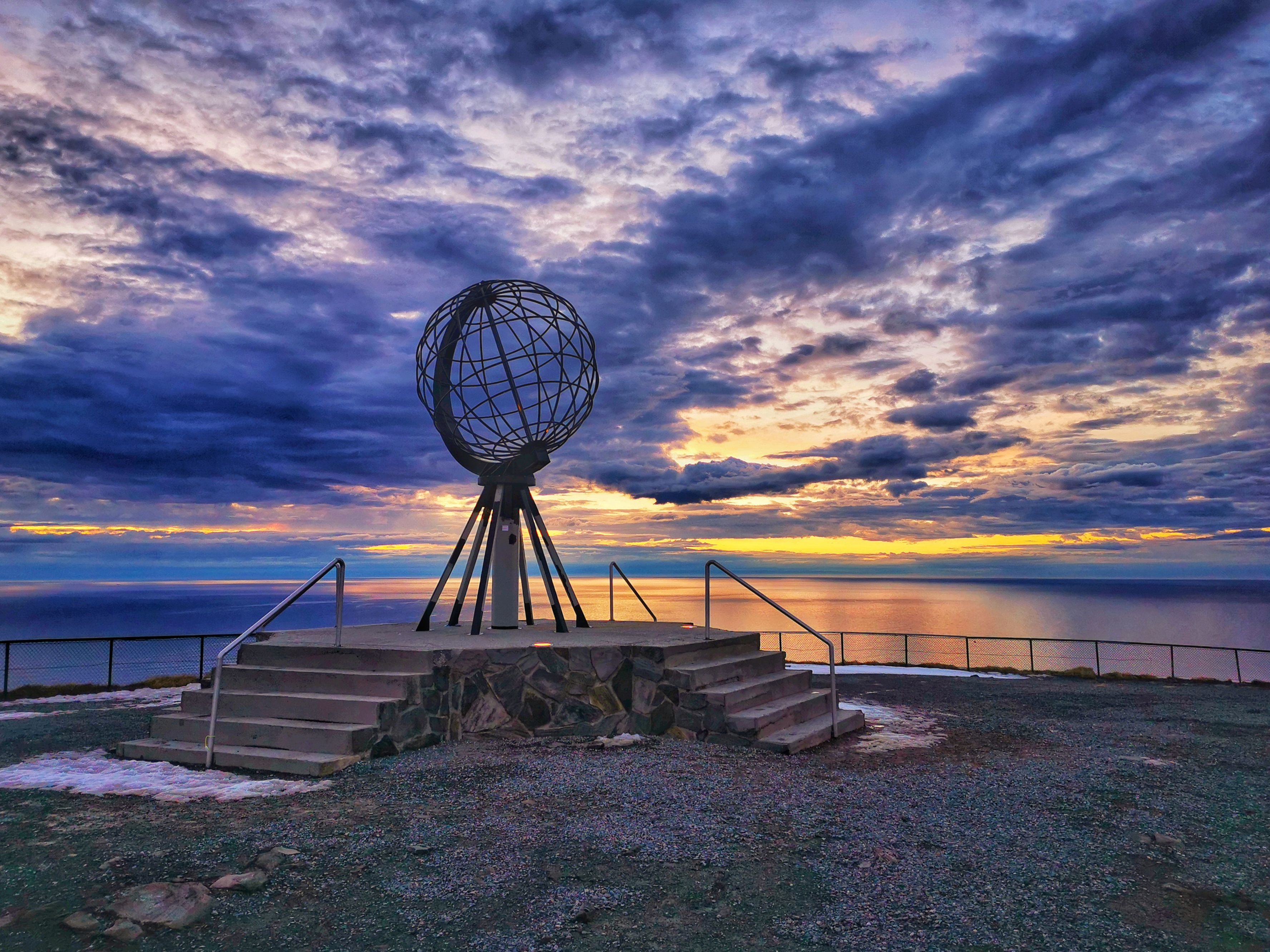 Die ikonische Nordkap-Kugel erhebt sich vor einem dramatischen arktischen Sonnenuntergang, während farbenprächtige Wolken sich über dem Barentssee spiegeln und einen atemberaubenden Blick auf Europas nördlichstes Wahrzeichen schaffen.
