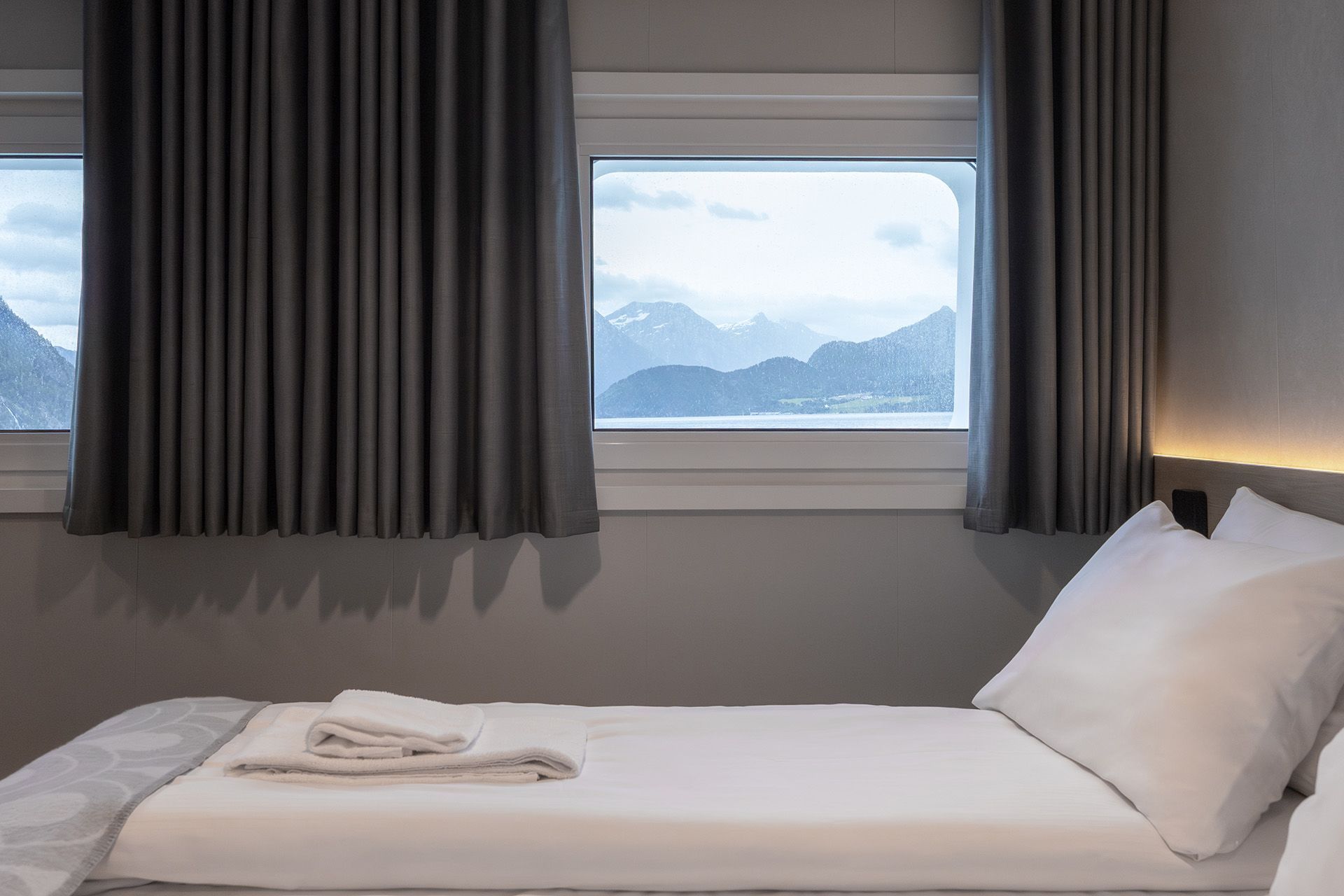 Aft View Superior Double cruise cabin with double bed and panoramic aft-facing window overlooking Norway’s coastal mountains.