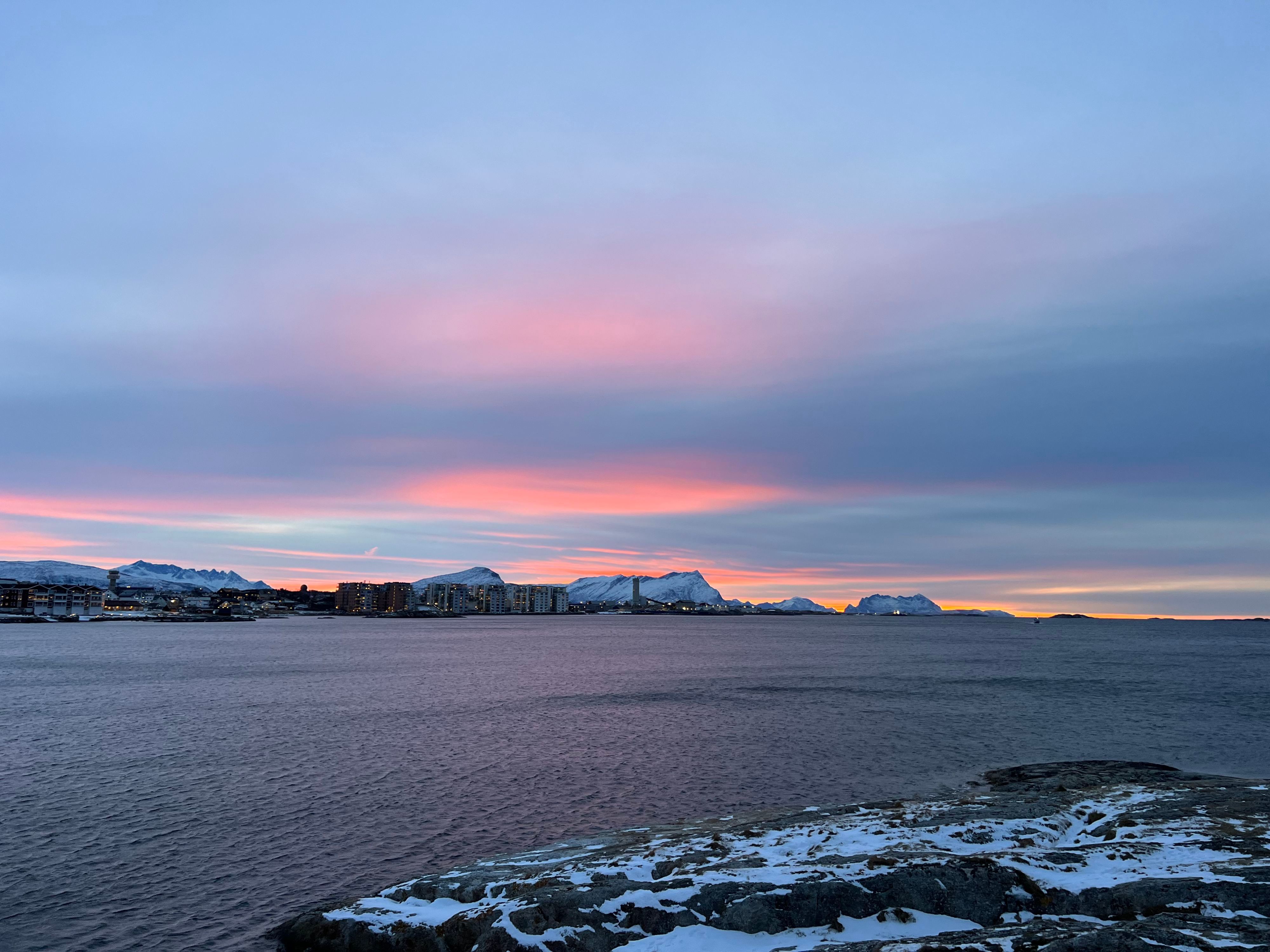 Vinter solnedgång över Bodøs strandpromenad med snötäckta berg och arktisk kustlinje.