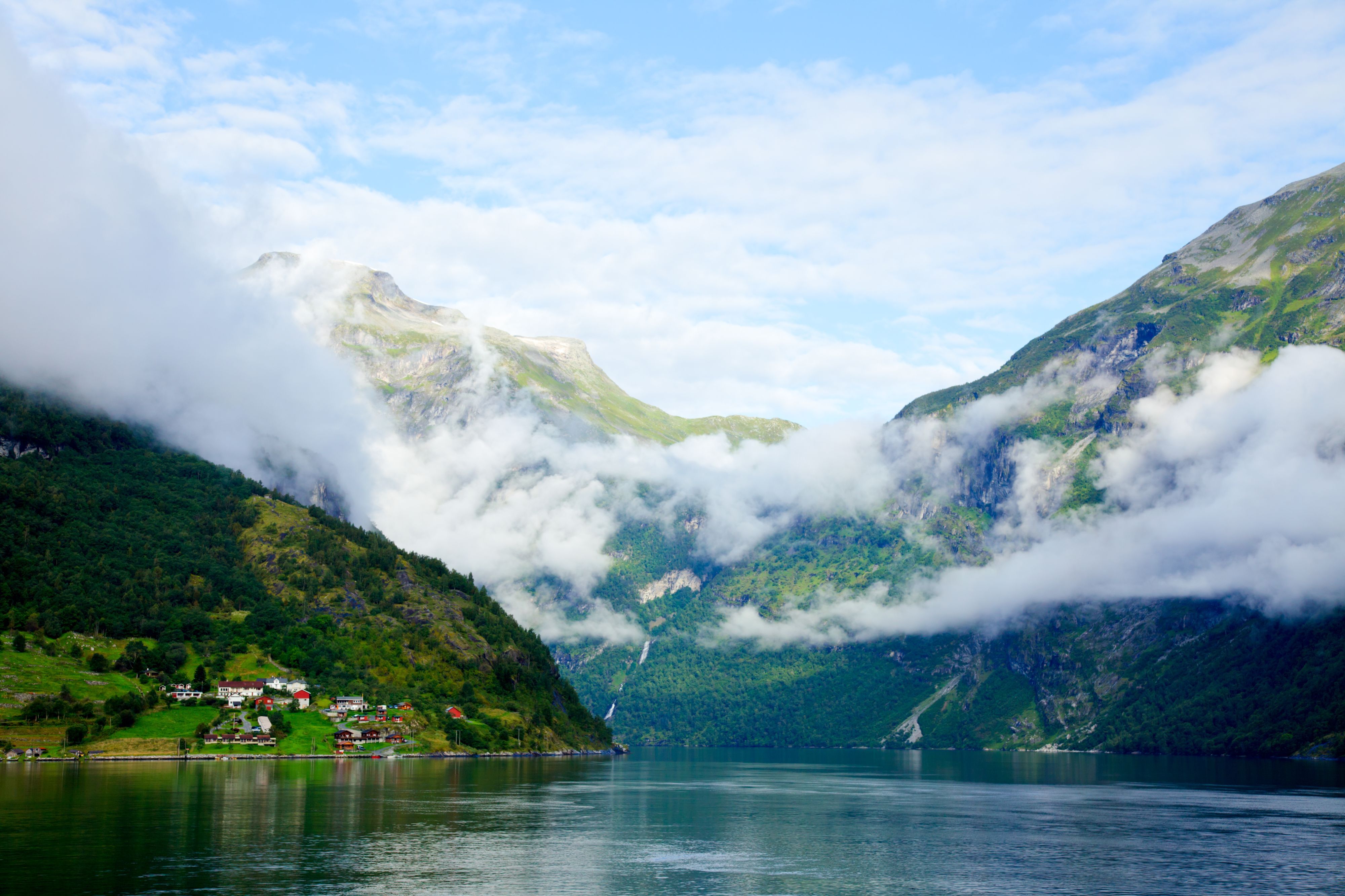 Lush green mountains wrapped in low clouds above the calm Geirangerfjord with a small village in spring (April), Norway